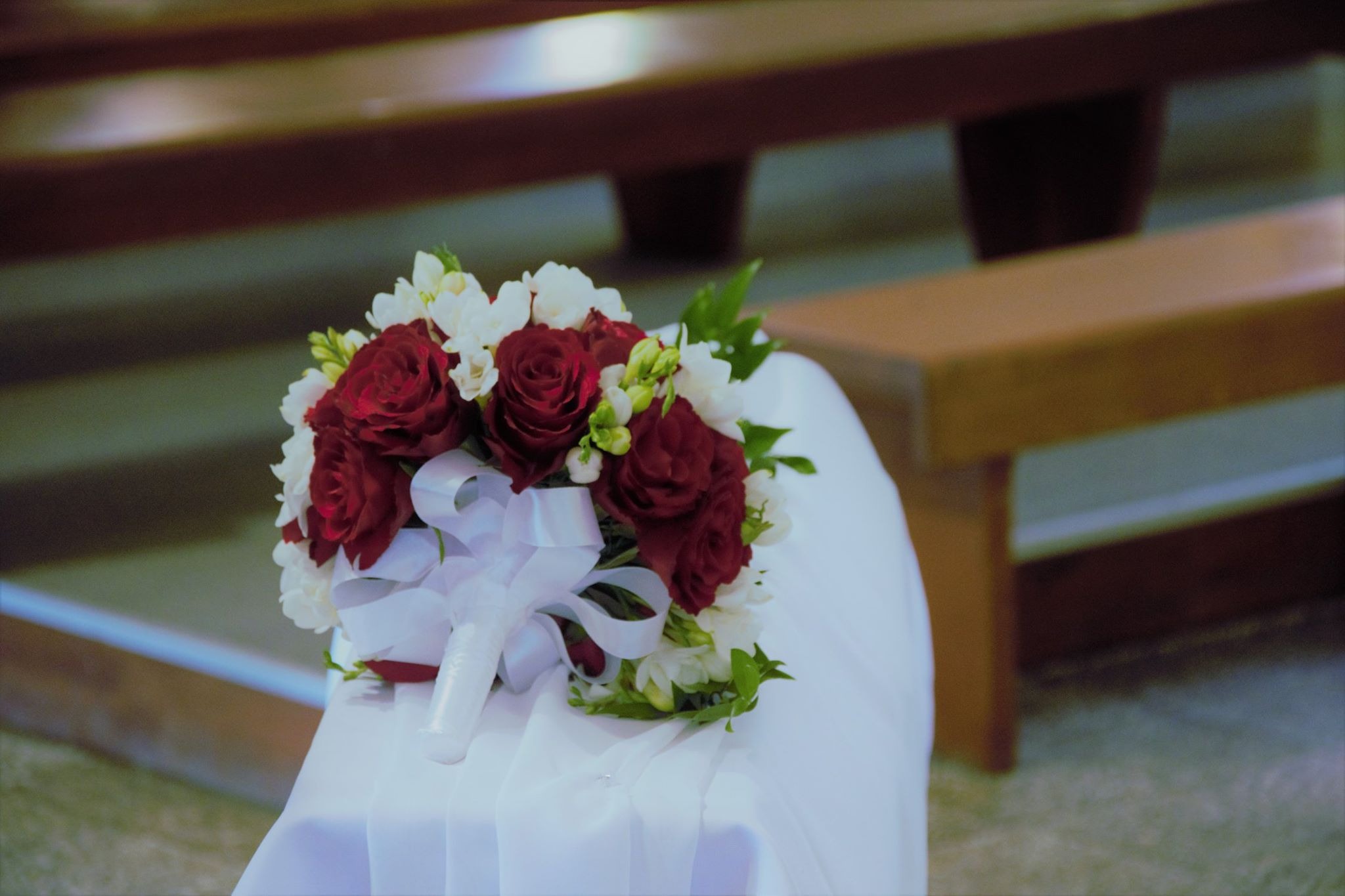 Bukiet ślubny z czerwonych róż i białych frezji, przewiązany białą wstążką, leży na białej tkaninie w kościele.