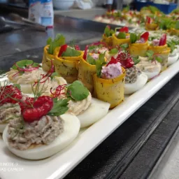 Elegancko podane przystawki: jajka faszerowane z dekoracją z malin i koperku oraz roladki z cukinii z farszem, na białym prostokątnym talerzu, na metalowym blacie.