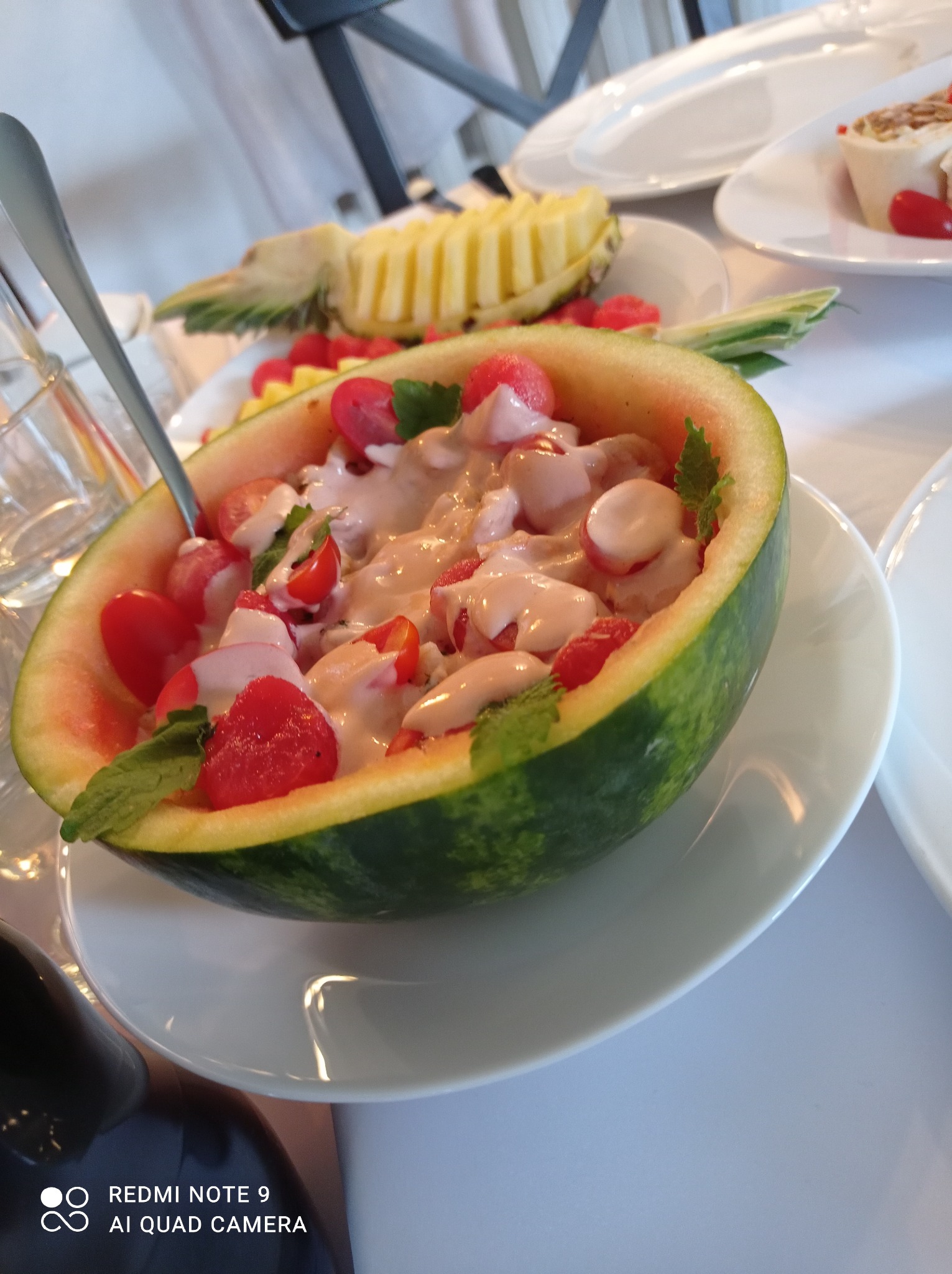 Arbuzowy półmisek z sałatką owocową polaną kremowym sosem, udekorowany listkami mięty, na białym talerzu, w tle ananas i przekąski.