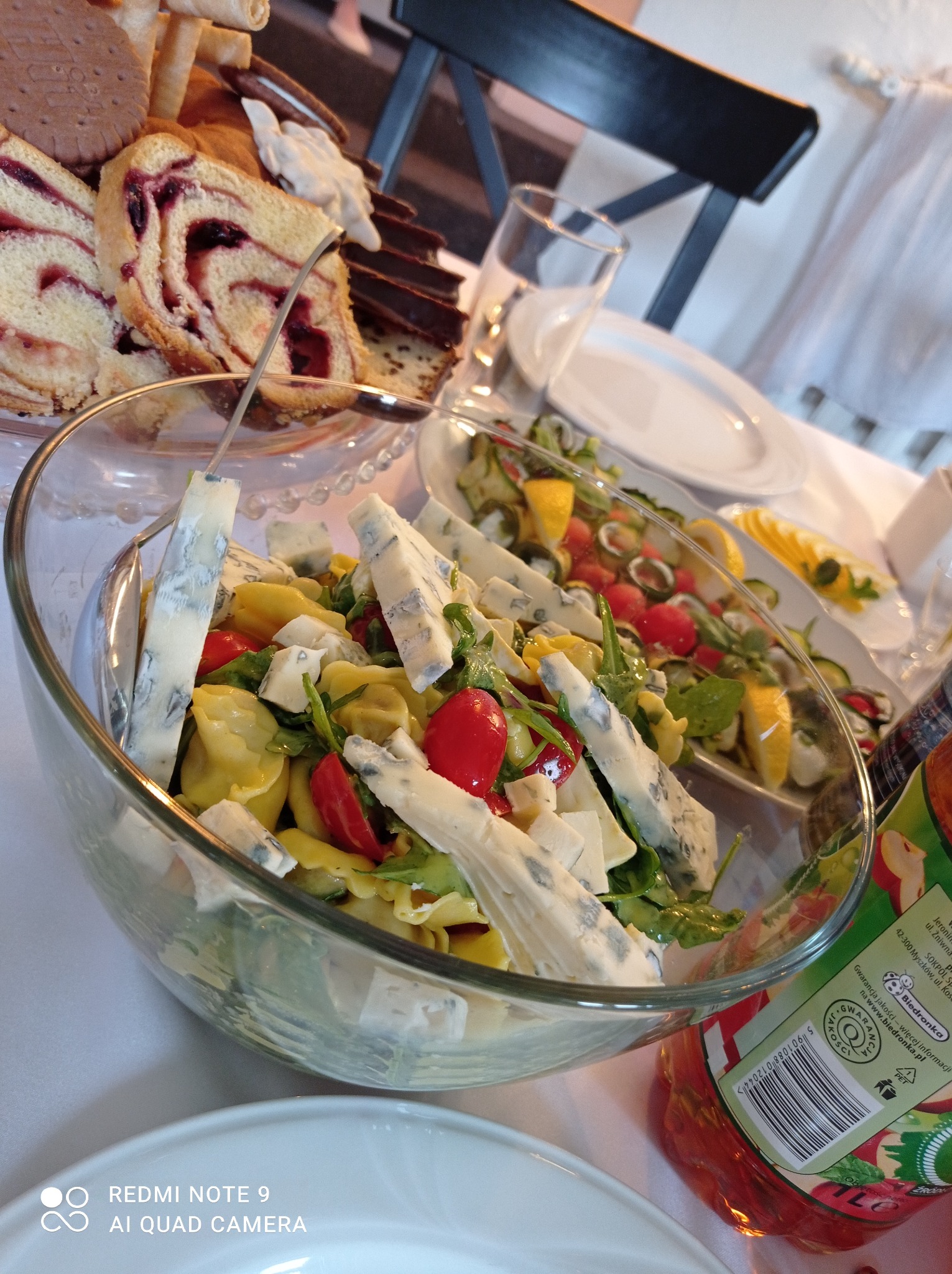 Szklana misa z sałatką tortellini, pomidorkami koktajlowymi, rukolą i kawałkami sera pleśniowego na stole z przekąskami, ciastami i napojami, przygotowana na przyjęcie.