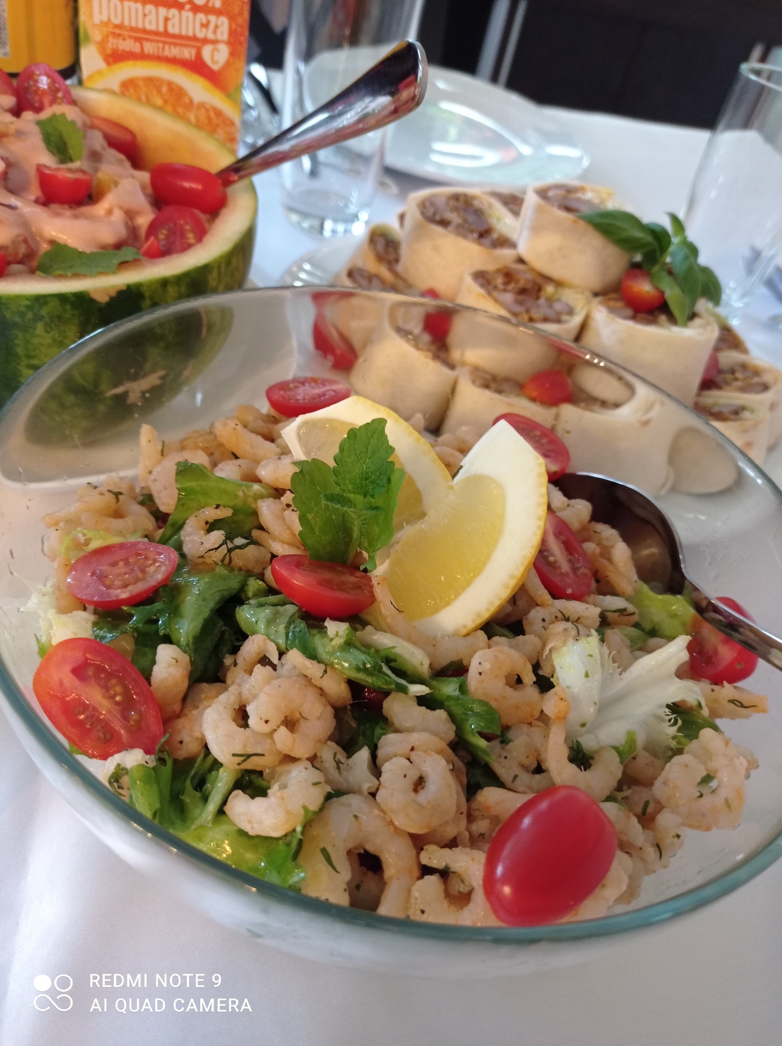 Szklana misa z sałatką krewetkową, pomidorkami cherry i cytryną, obok roladki z farszem i arbuz wypełniony sałatką z sosem pomarańczowym, na białym obrusie.