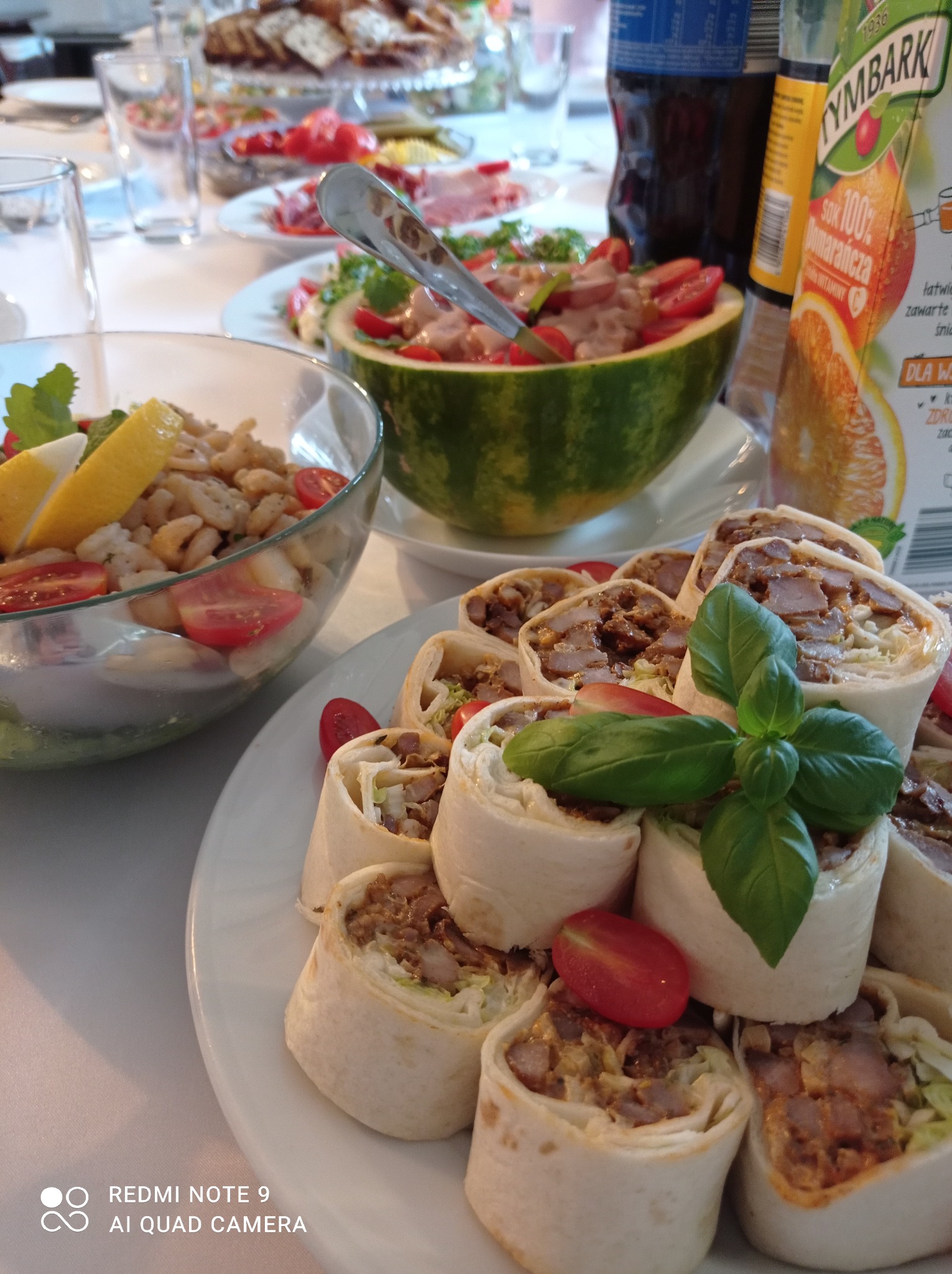 Stół z różnorodnymi przekąskami: roladki z tortilli z mięsem, sałatka z fasolą i pomidorami, sałatka w wydrążonym arbuzie, napoje w tle.