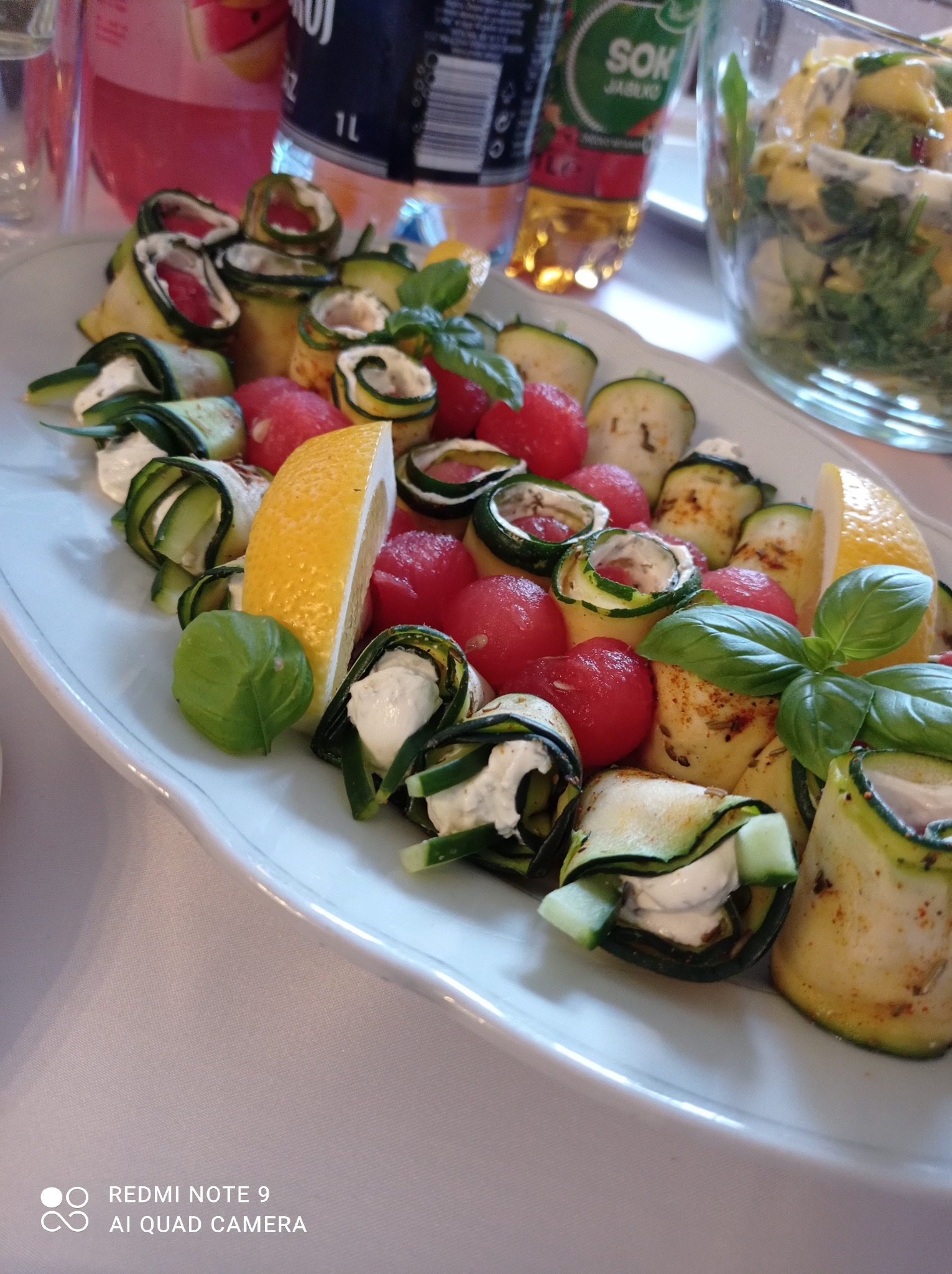 Elegancka kompozycja na białym talerzu: roladki z grillowanej cukinii z białym serem, przeplatane kulkami arbuza i plastrami cytryny, ozdobione świeżą bazylią, z napojami w tle, sugerująca catering...