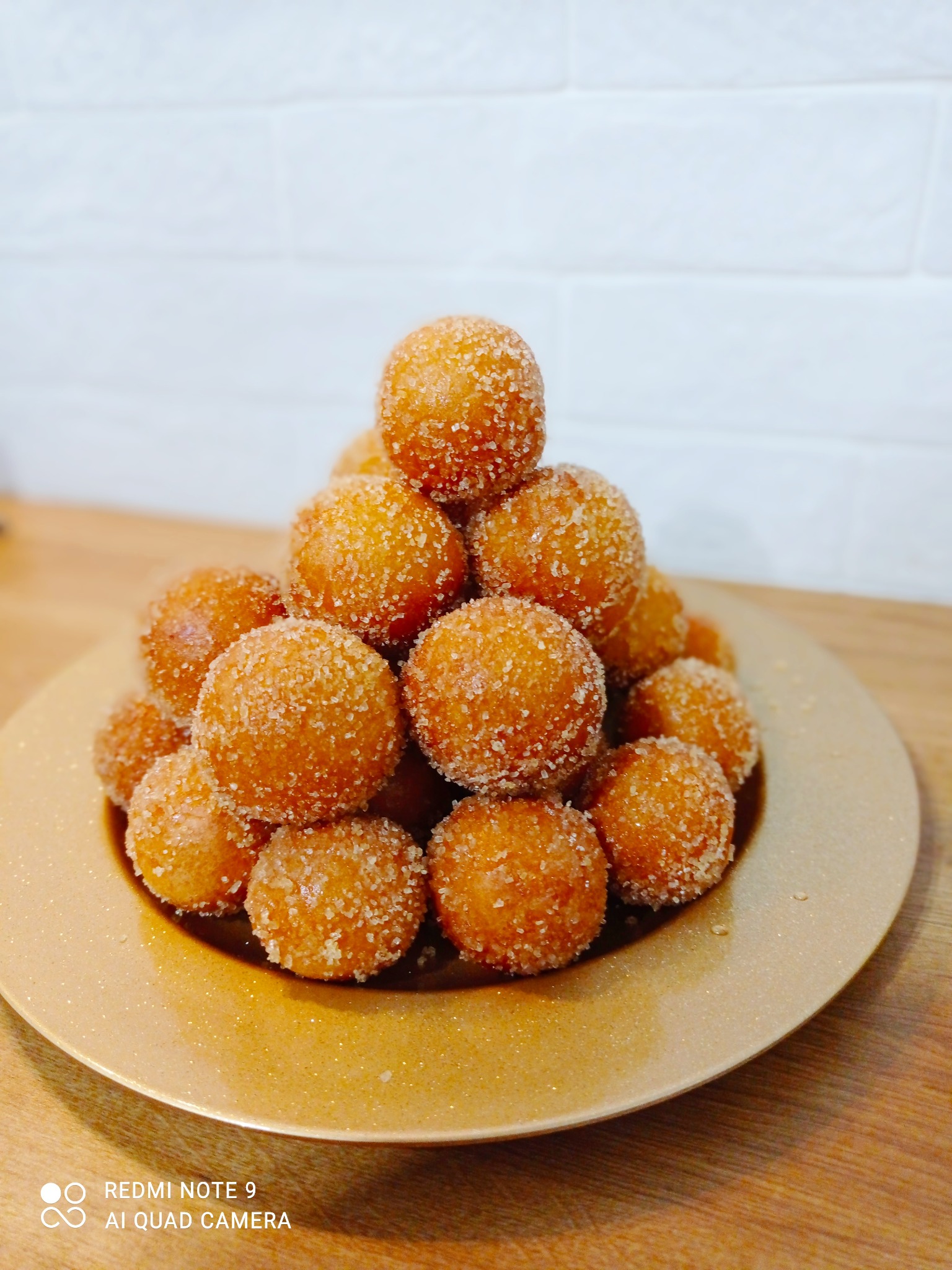 Kopiec pączków w cukrze pudrze ułożonych na złotej tacy, na drewnianym blacie i tle z białej cegły.