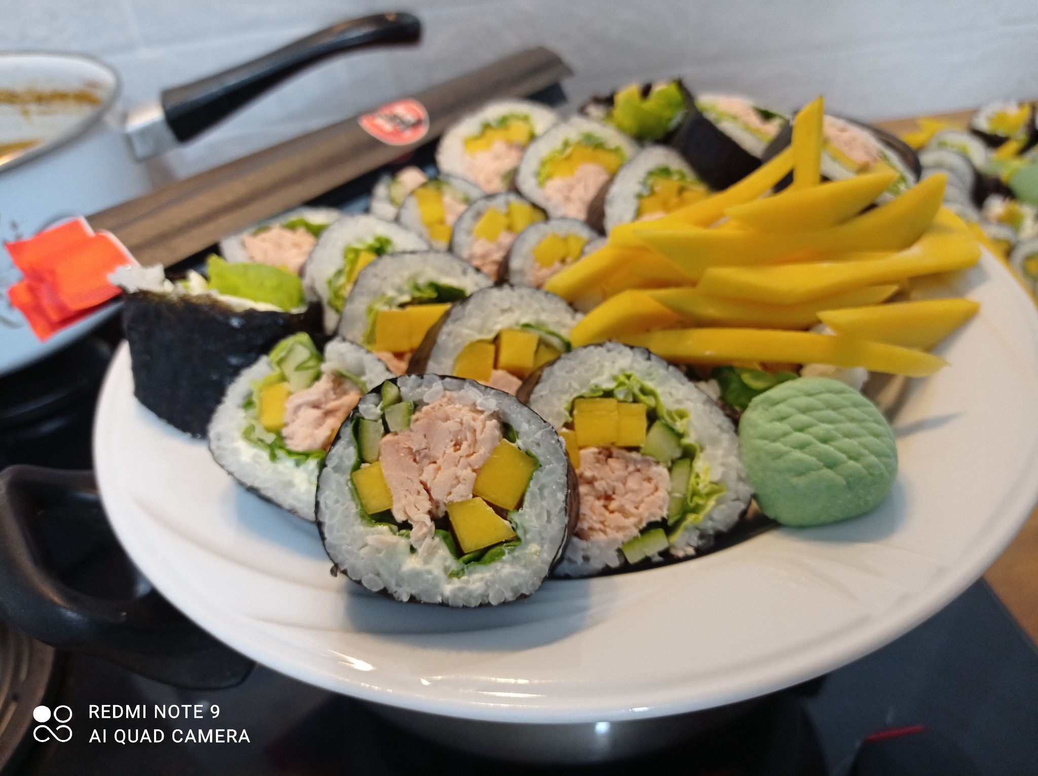 Talerz z sushi: rolki z łososiem, mango i ogórkiem, podane z wasabi; w tle garnek i łopatka kuchenna.