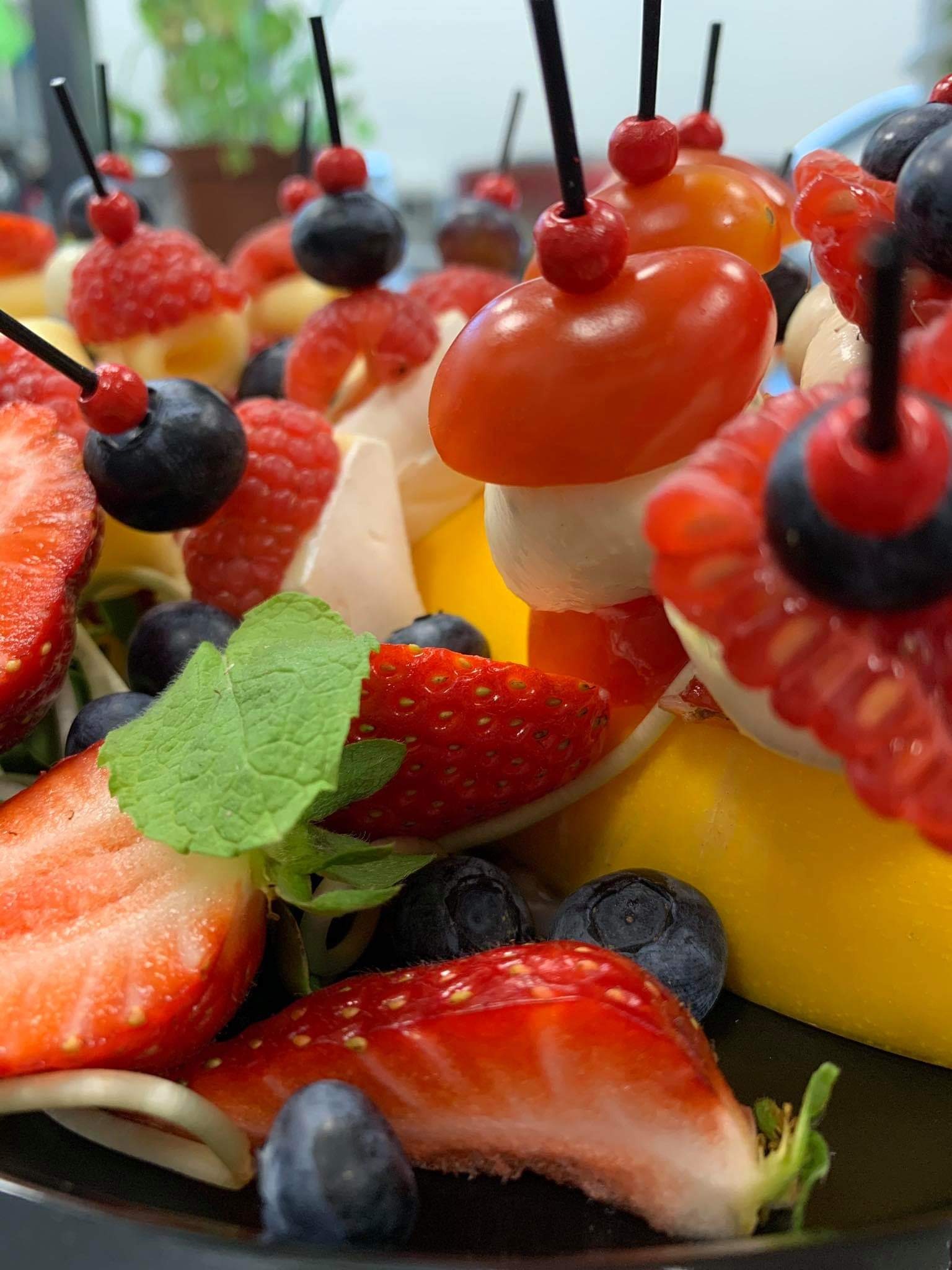 Zbliżenie na kolorowe koreczki owocowo-warzywne: truskawki, borówki, maliny, pomidorki cherry i mini kulki mozzarelli, spięte wykałaczkami, ułożone na czarnym talerzu.