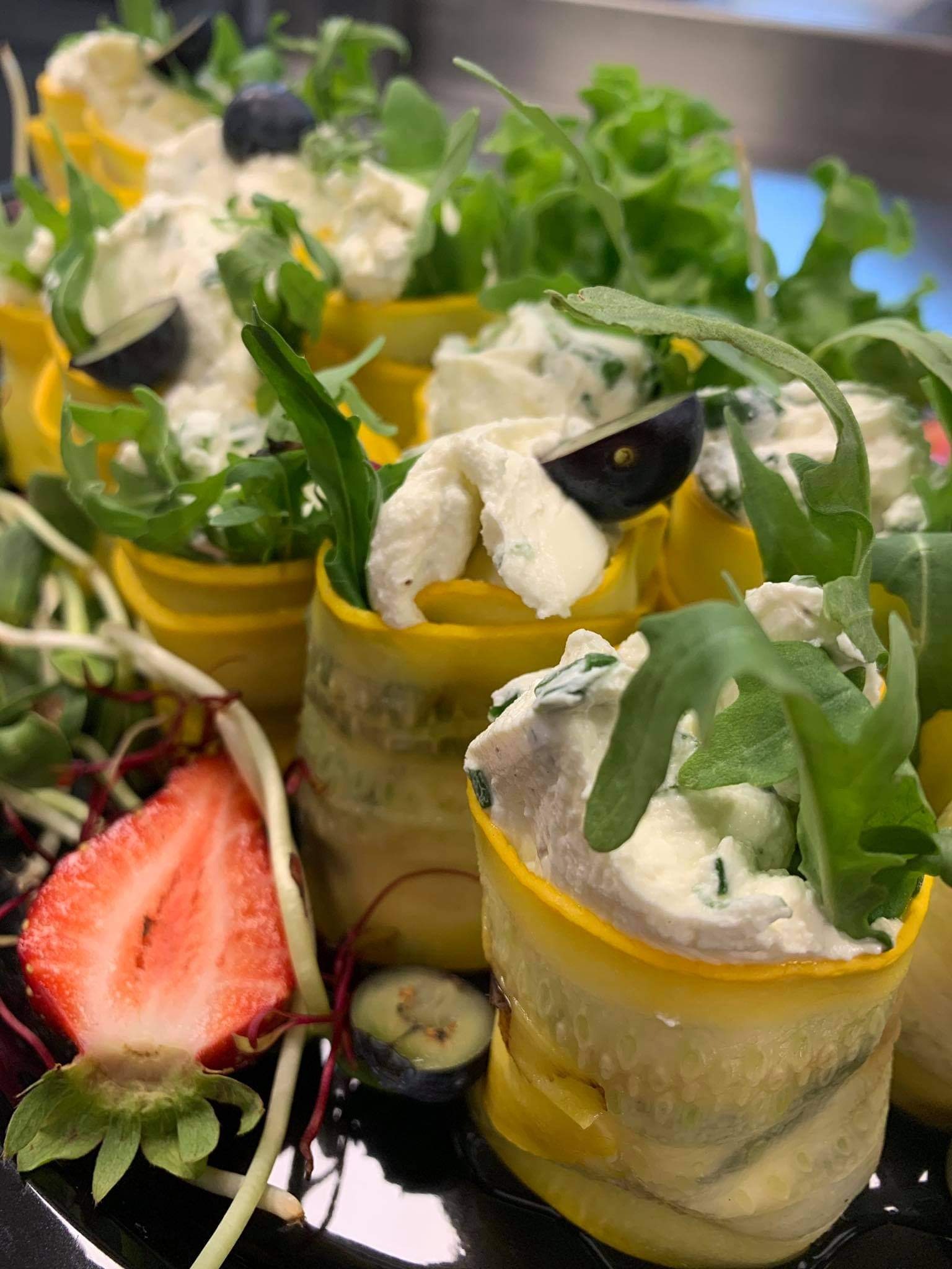 Żółte roladki z cukinii wypełnione kremowym serem i rukolą, udekorowane borówkami i połówką truskawki na czarnym talerzu.