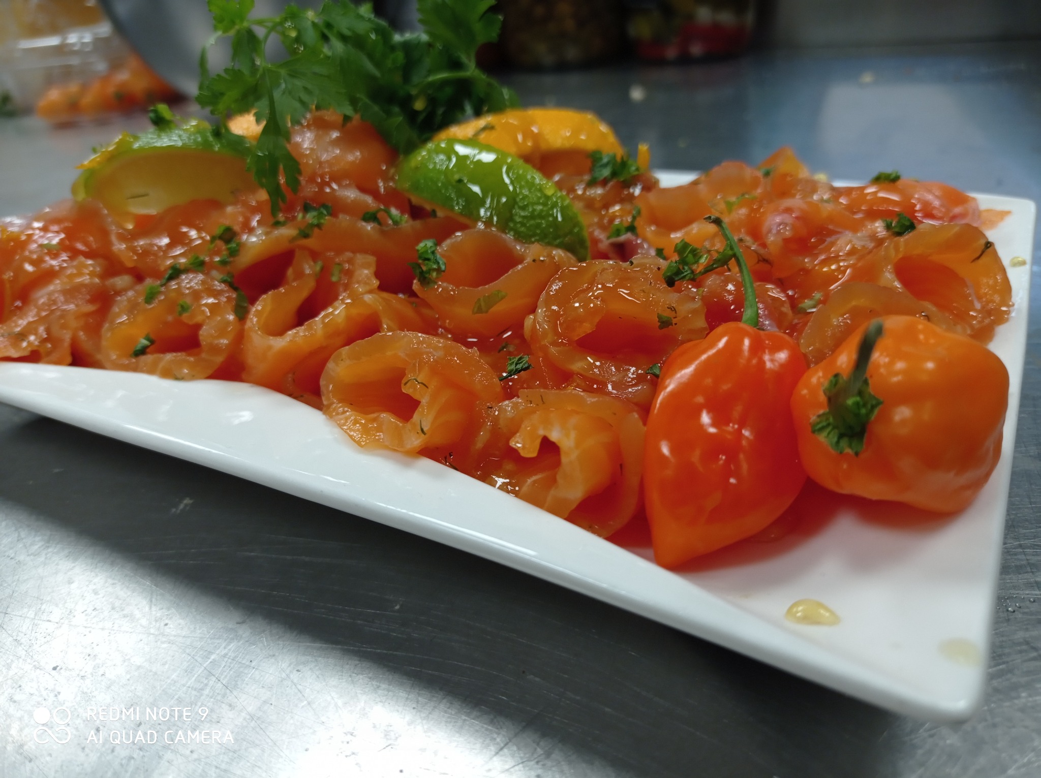 Carpaccio z łososia ułożone na białym talerzu, udekorowane świeżymi ziołami, plasterkami limonki i pomarańczowej papryki habanero, na metalowym blacie kuchennym.