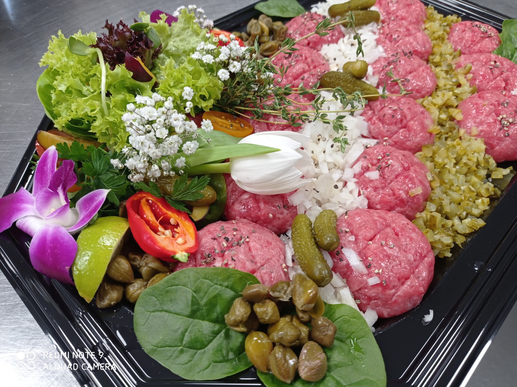 Elegancko ułożona taca z tatarem wołowym, dekorowana sałatą, kaparami, ogórkami konserwowymi, cebulą, kwiatami i ziołami.