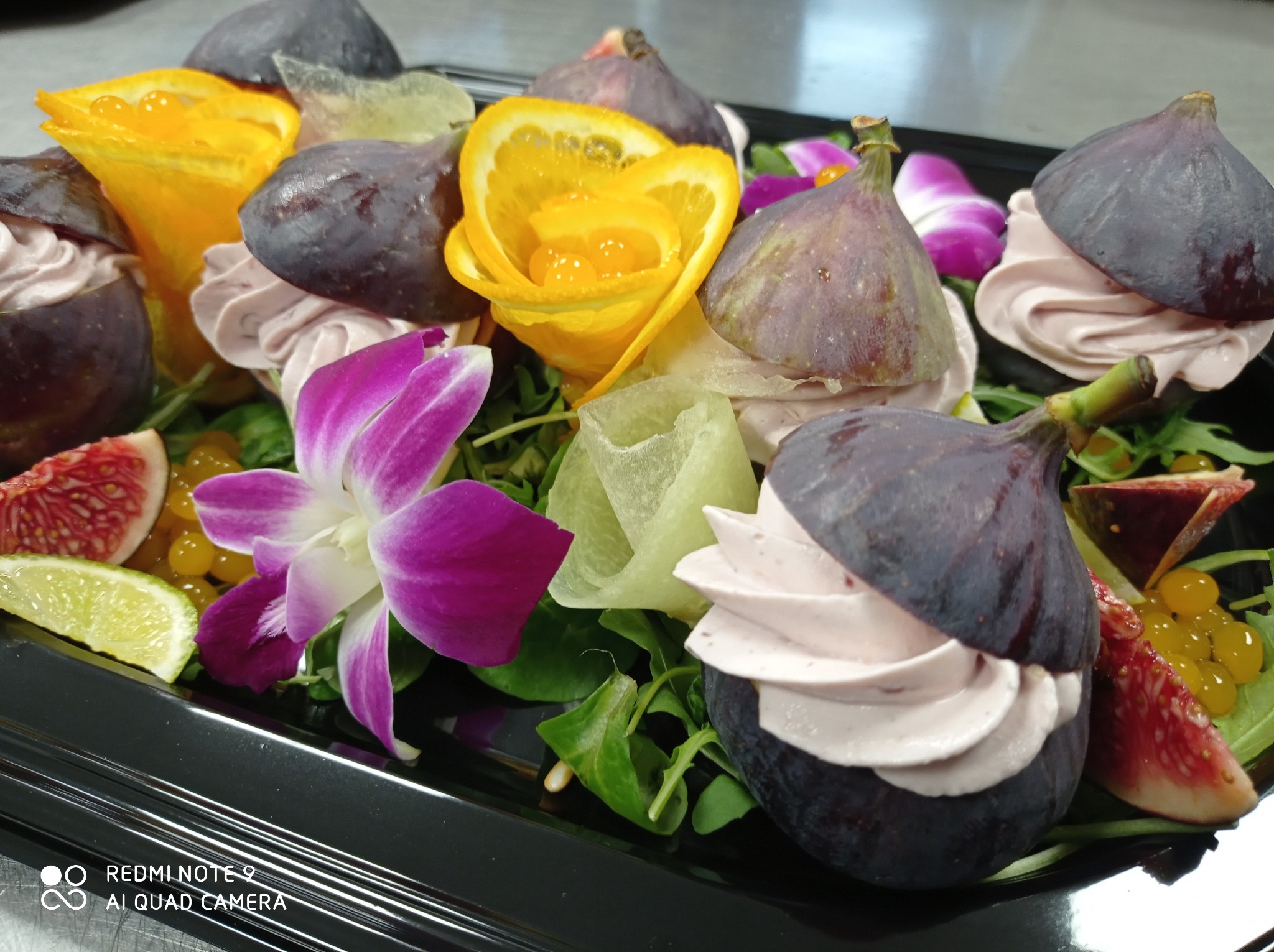 Elegancka kompozycja cateringowa: figi z kremem, dekoracje z pomarańczy i kwiatów na czarnej tacy.