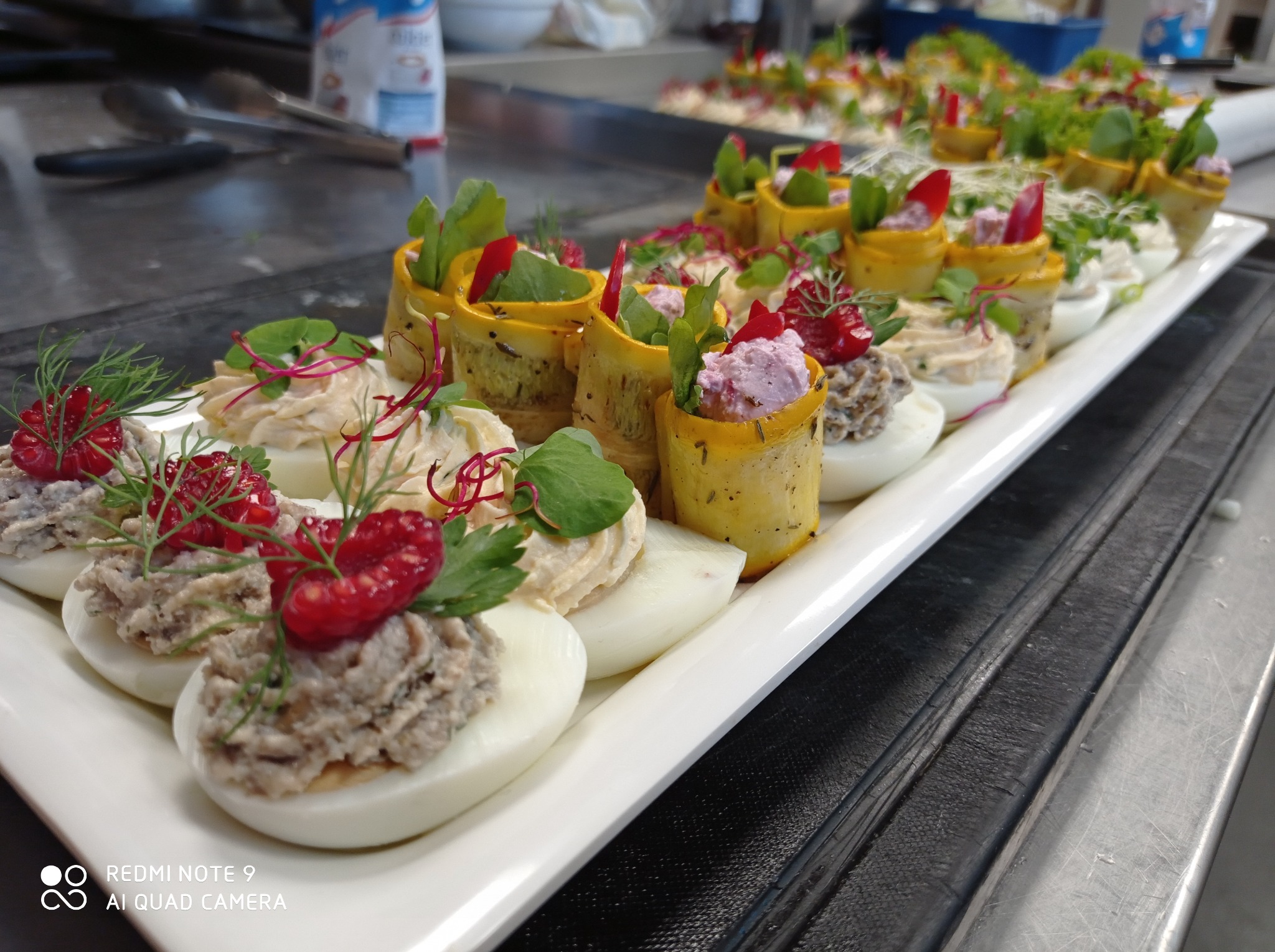 Elegancko podane przystawki: jajka faszerowane z dekoracją z malin i koperku oraz roladki z cukinii z farszem, na białym prostokątnym talerzu, na metalowym blacie.