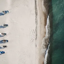 Widok z góry na plażę z piaskiem i turkusową wodą, wzdłuż brzegu ułożonych jest pięć łodzi z różnymi kolorami kadłubów, pozostawiając ślady na piasku.