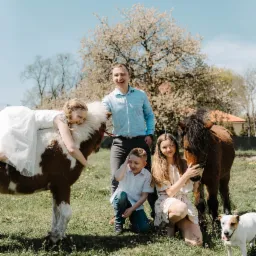 Uśmiechnięta rodzina z dwójką dzieci i psem pozuje z dwoma małymi końmi na łące w słoneczny dzień, kwitnące drzewo w tle.
