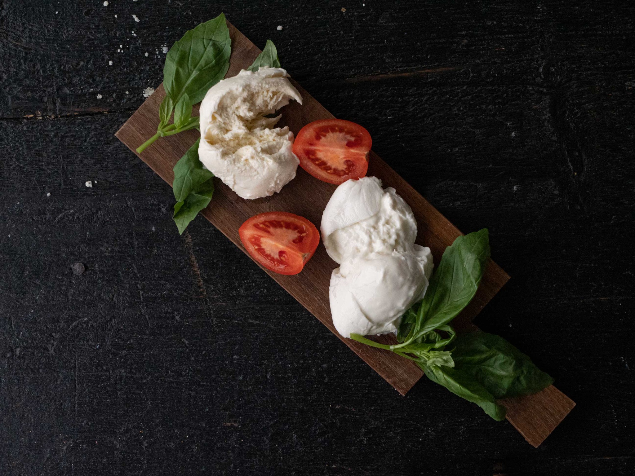 Deska z dwoma kulkami mozzarelli, połówkami pomidora i świeżą bazylią na ciemnym, drewnianym tle z posypką przypominającą sól.
