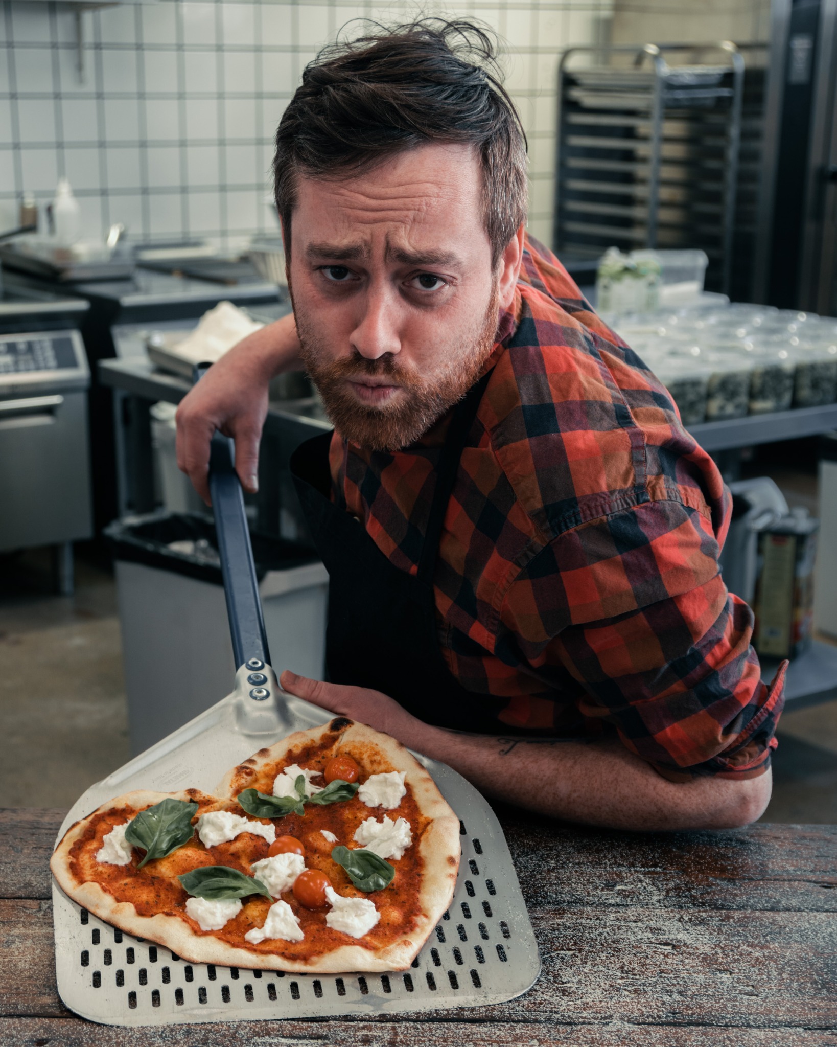 Kucharz w kuchni prezentuje pizzę w kształcie serca na metalowej łopacie, z pomidorami, bazylią i serem mozzarella, spoglądając wprost w obiektyw z lekkim niedowierzaniem.