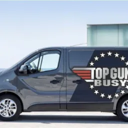 Szary van z logo firmy transportowej 'TOP GUN BUSY' na tle nowoczesnego budynku i morza.