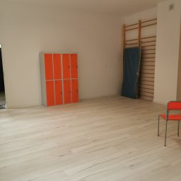 Firma Ogólnobudowlana SSWG - Jasne pomieszczenie z pomarańczowymi szafkami, drabinkami gimnastycznymi z materacem i krzesłem. Podłoga wyłożona panelami imitującymi drewno, oświetlenie sufitowe.