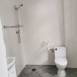Firma Ogólnobudowlana SSWG - Nowoczesna, jasna łazienka z białymi płytkami ściennymi, szarą podłogą w płytkach, toaletą z uchwytem dla niepełnosprawnych i chromowaną armaturą prysznicową.