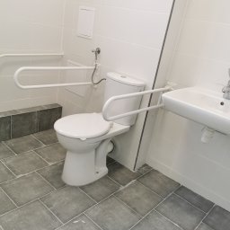 Firma Ogólnobudowlana SSWG - Biała toaleta z uchwytami dla osób niepełnosprawnych zamontowanymi na ścianie z białych płytek, szara podłoga z płytek, biała umywalka.