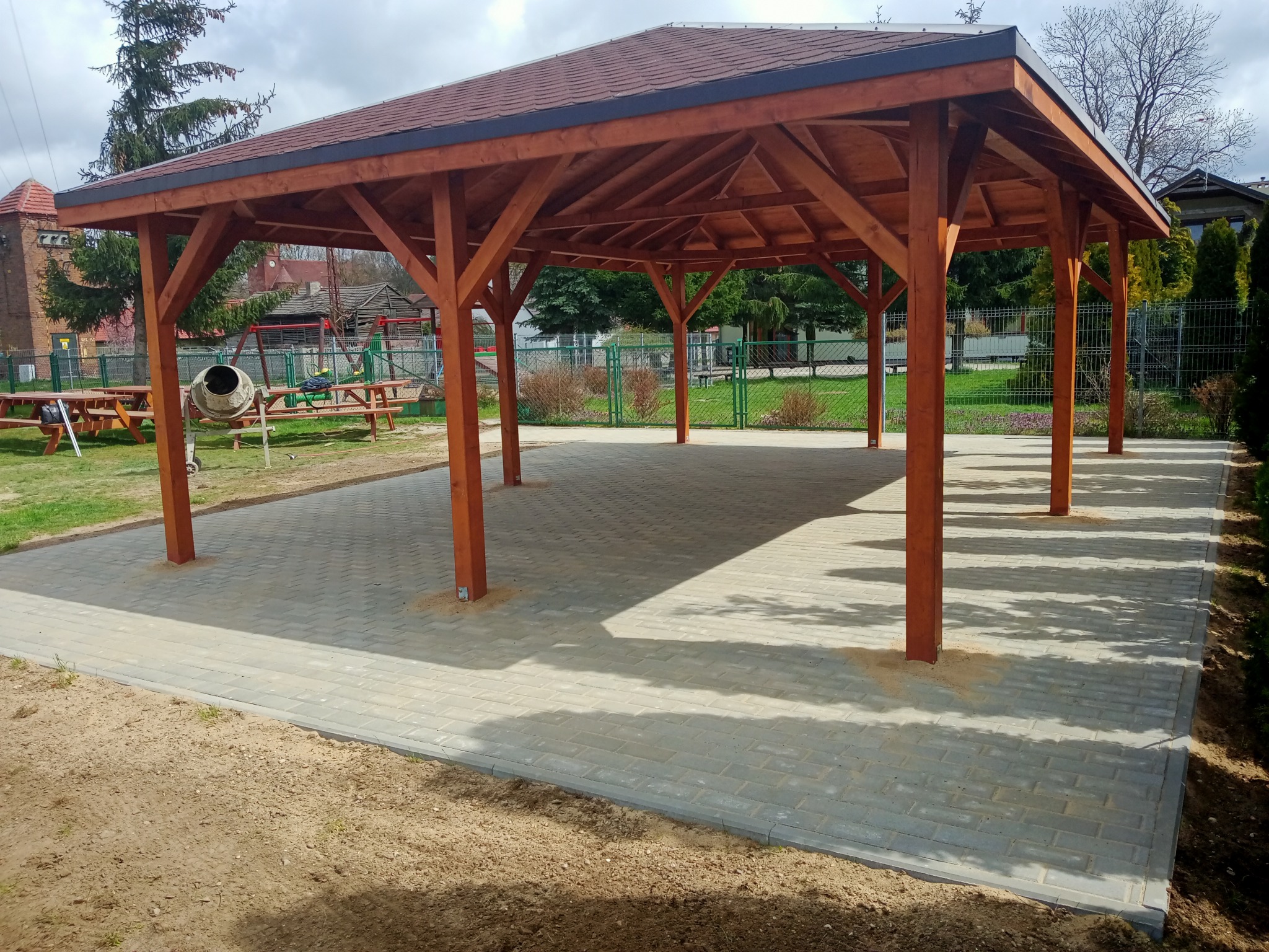 Drewniana altana z brązowym dachem, stojąca na wybrukowanym placu z kostki, z widoczną betoniarką w tle.