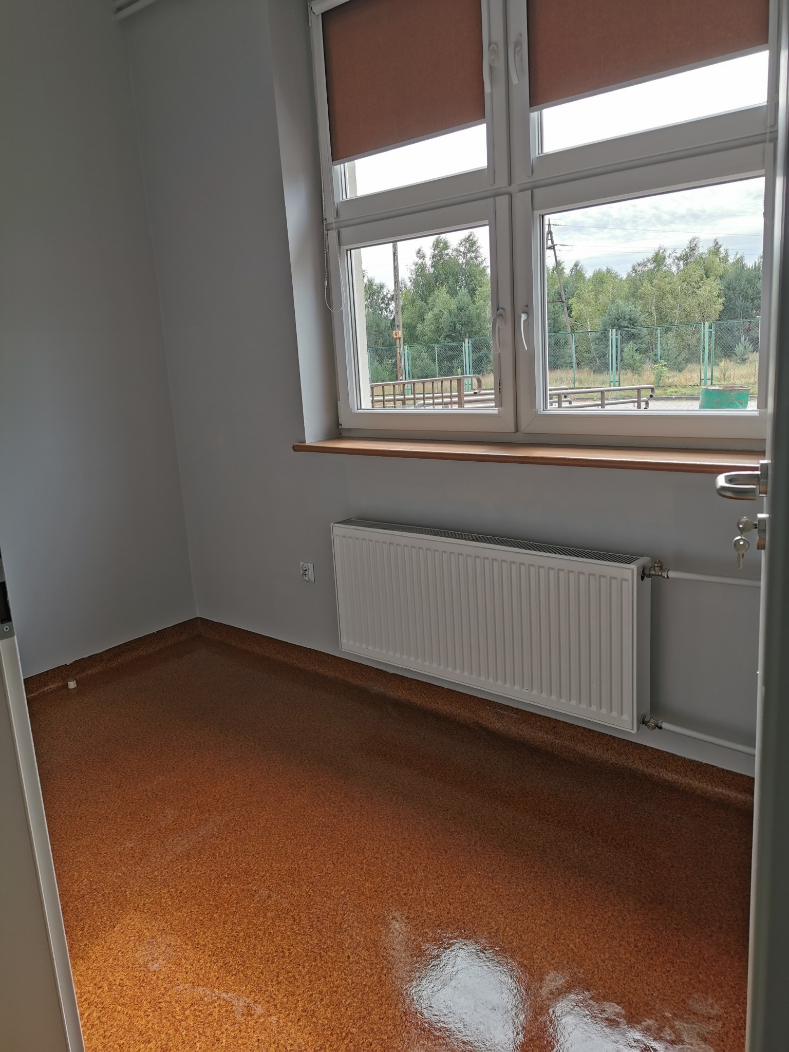 Pomalowane na szaro pomieszczenie z brązową podłogą, białym grzejnikiem pod oknem z brązowymi roletami.