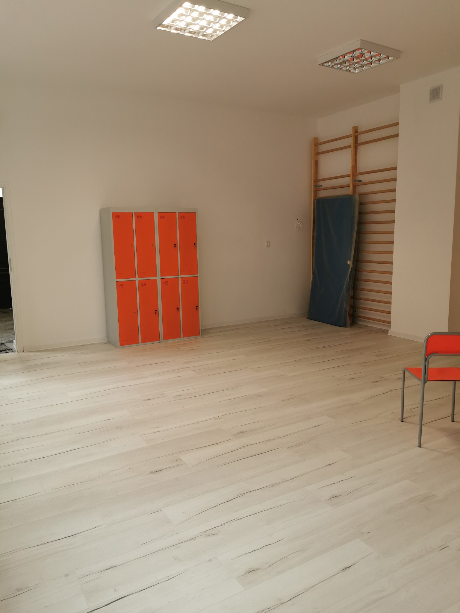 Jasne pomieszczenie z pomarańczowymi szafkami, drabinkami gimnastycznymi z materacem i krzesłem. Podłoga wyłożona panelami imitującymi drewno, oświetlenie sufitowe.