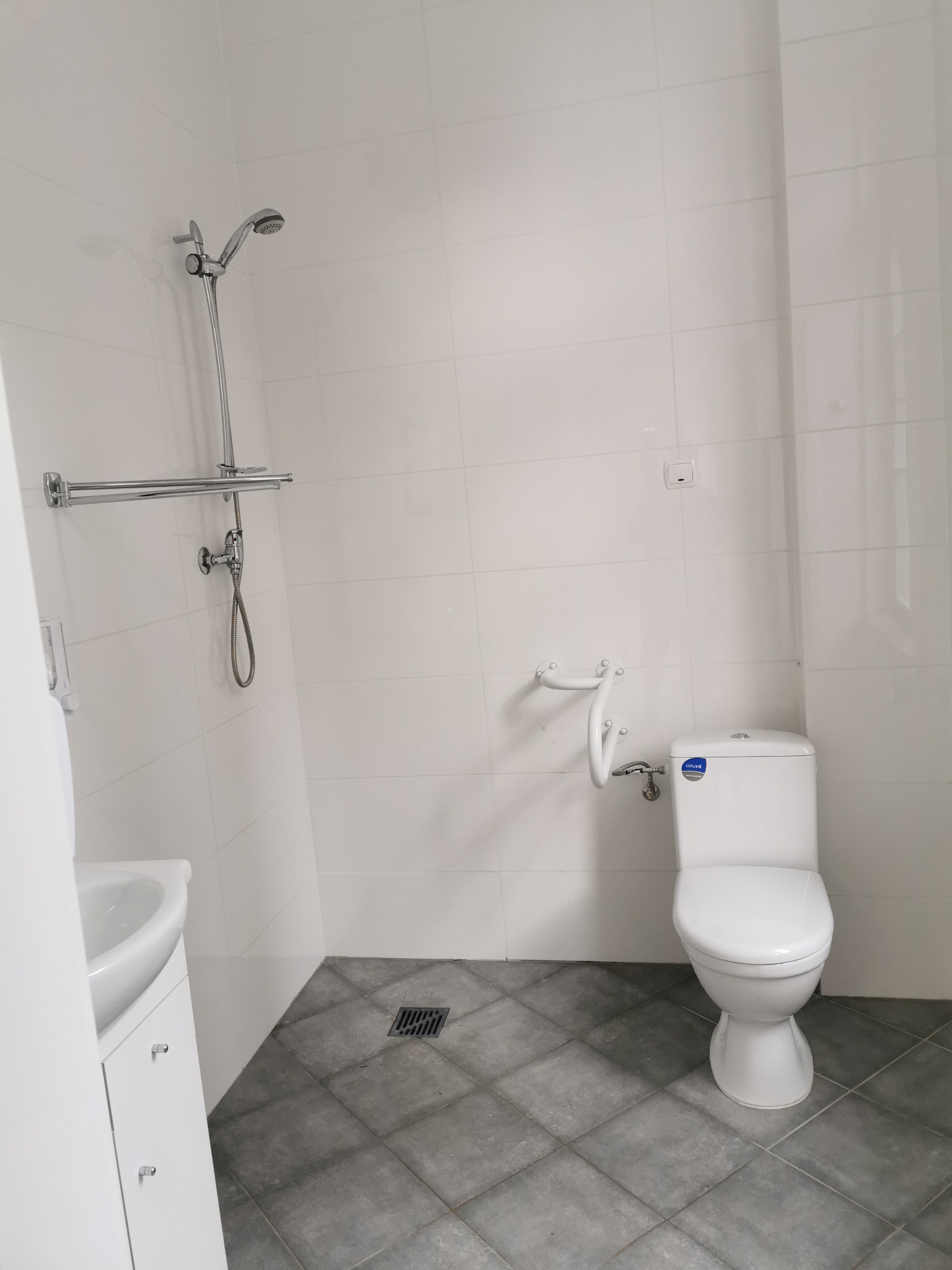 Nowoczesna, jasna łazienka z białymi płytkami ściennymi, szarą podłogą w płytkach, toaletą z uchwytem dla niepełnosprawnych i chromowaną armaturą prysznicową.