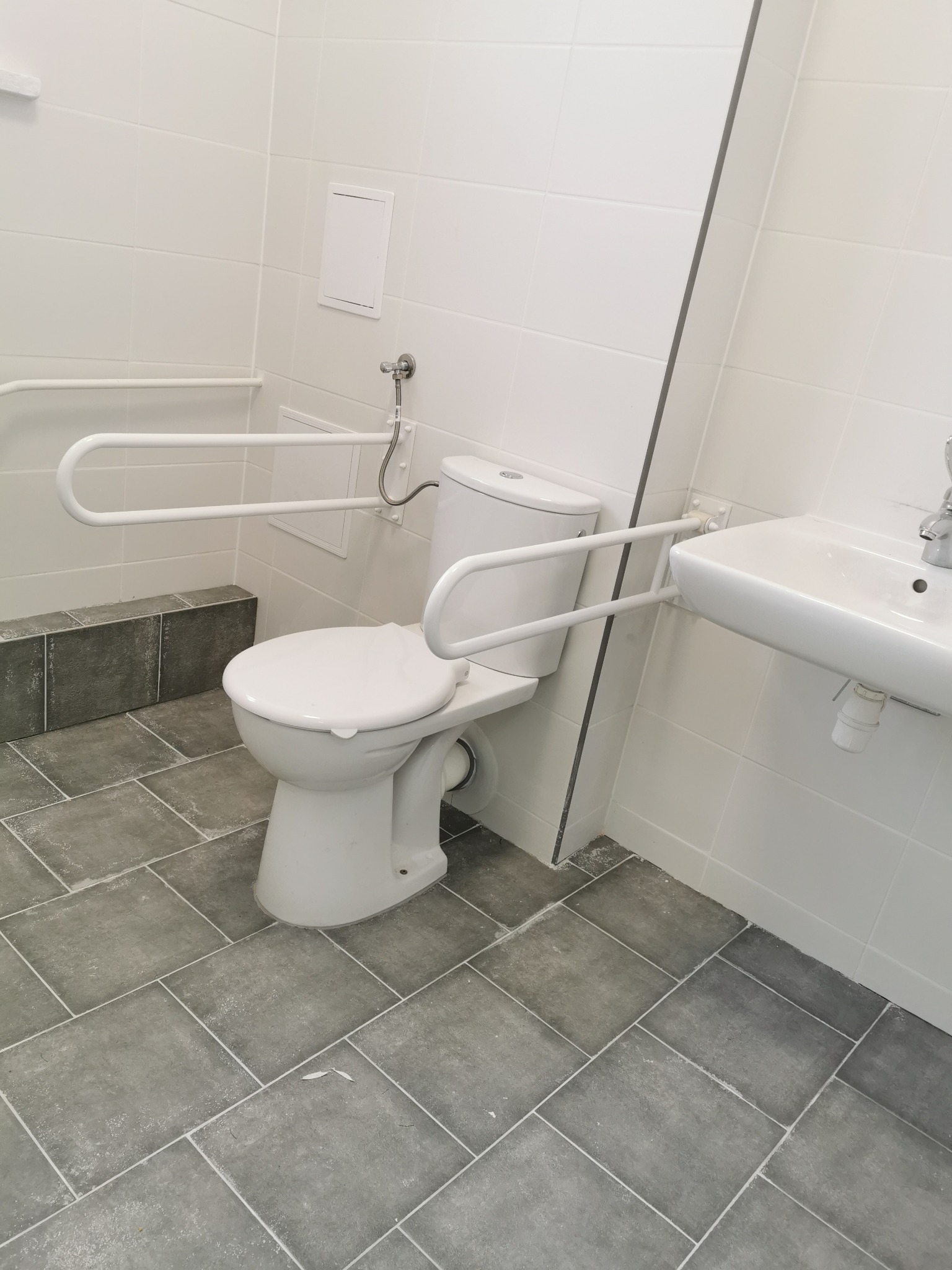 Biała toaleta z uchwytami dla osób niepełnosprawnych zamontowanymi na ścianie z białych płytek, szara podłoga z płytek, biała umywalka.