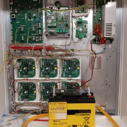 Otwarta szafa sterownicza z elektroniką alarmową, liczne płytki drukowane, kable i akumulator 12V Sunbattery SB12-18 z ostrzeżeniem w języku niemieckim i angielskim o konieczności ładowania przez...