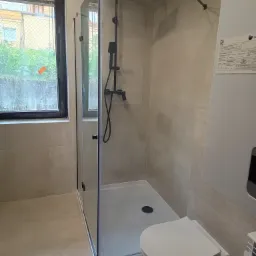 Wykończona łazienka z kabiną prysznicową typu walk-in, podwieszaną toaletą i beżowymi płytkami. Widoczne czarne elementy armatury i okno z widokiem na zieleń.