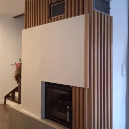 Zabudowa kominkowa z pionowymi drewnianymi listwami i gładką, białą płytą, pod którą znajduje się czarny wkład kominkowy z szybą. Podstawa kominka wykończona szarą płytką imitującą kamień. Widoczny...