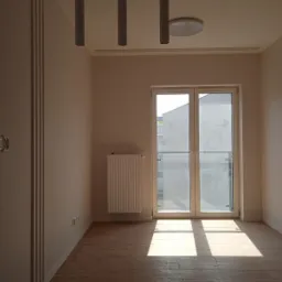 Pusty pokój w stanie deweloperskim z panelami podłogowymi, grzejnikiem, białymi ścianami, trzema cylindrycznymi lampami wiszącymi i drzwiami balkonowymi wpuszczającymi silne światło słoneczne.