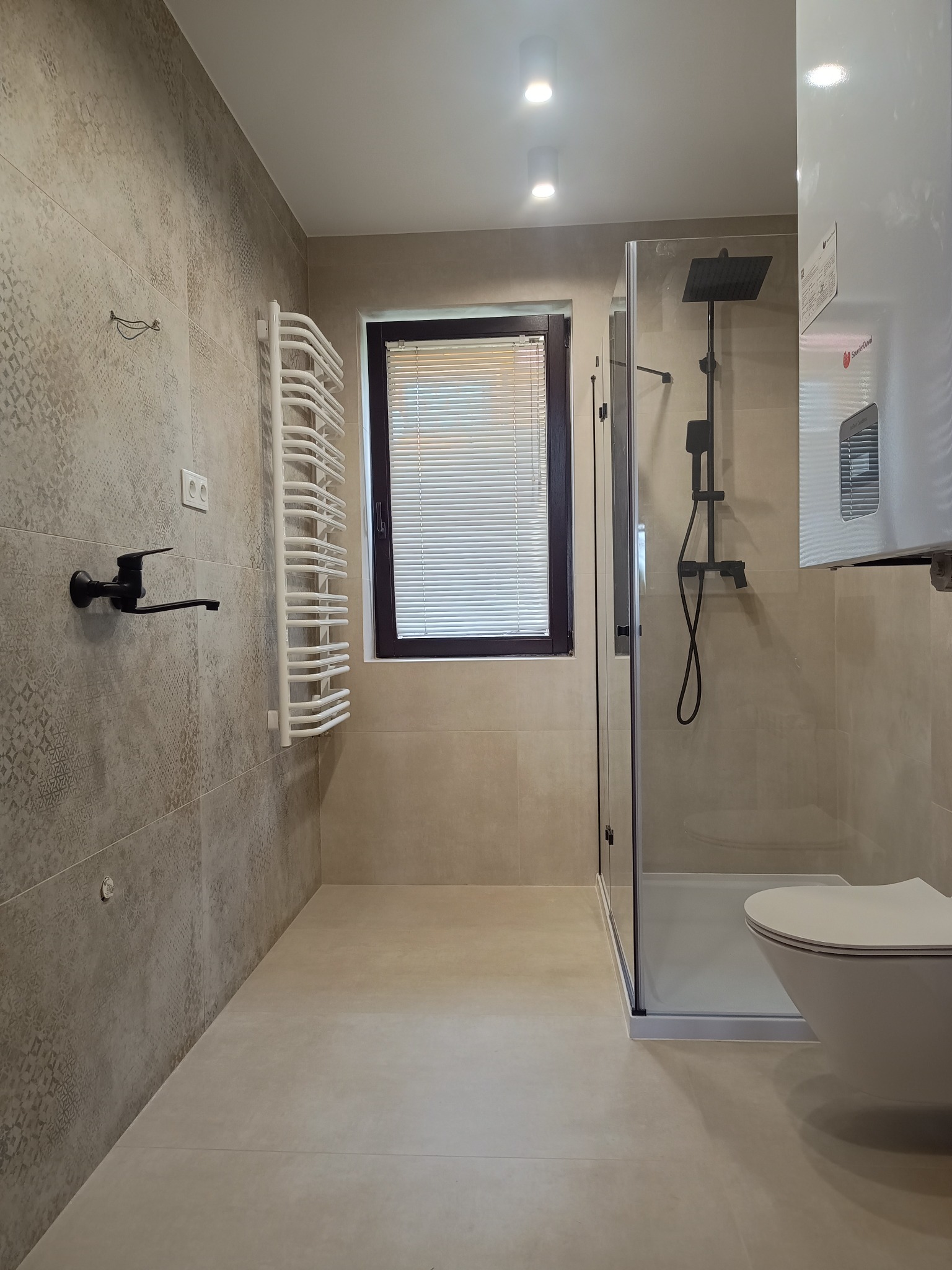 Jasna łazienka po remoncie: beżowe płytki, czarna armatura, kabina prysznicowa z deszczownicą i grzejnik drabinkowy. Minimalistyczny design, okno z żaluzjami.