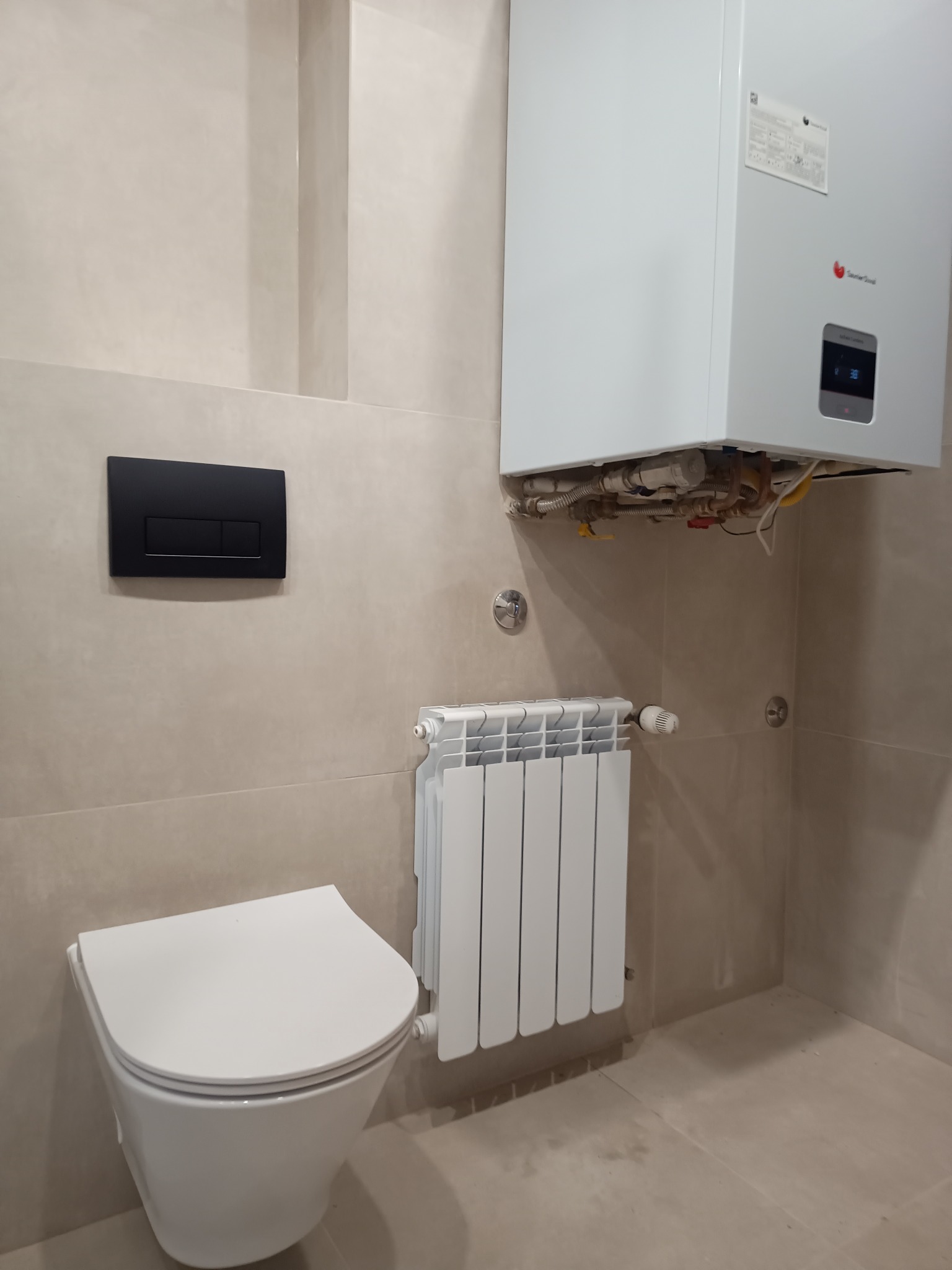 Wykończona łazienka w beżach: wisząca toaleta, biały grzejnik i piec gazowy. Minimalistyczny design, płytki imitujące kamień, czarny panel spłuczki.