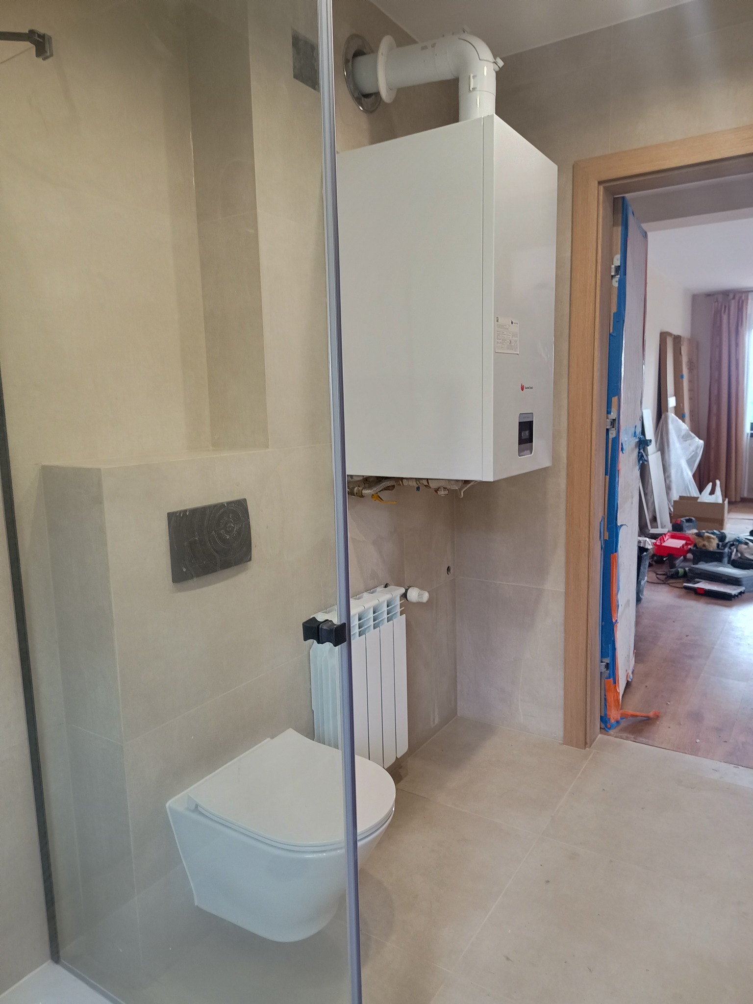 Wykończona łazienka z beżowymi płytkami, wiszącą toaletą i grzejnikiem. Na ścianie zamontowany piec gazowy. Widoczna szklana kabina prysznicowa i otwarte drzwi.