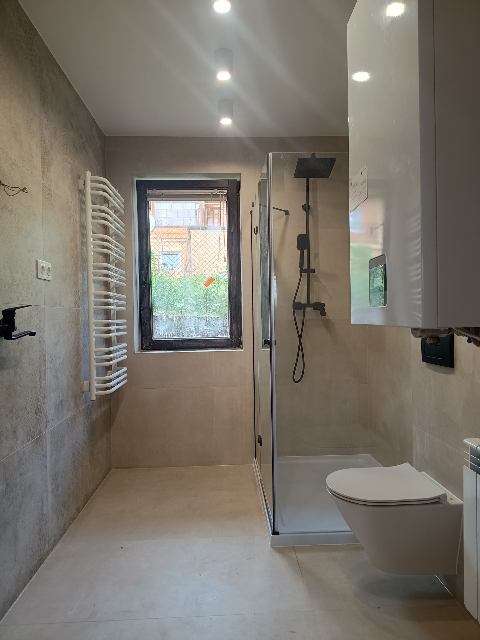 Jasna łazienka po remoncie z kabiną prysznicową, podwieszaną toaletą i grzejnikiem drabinkowym. Beżowe płytki na ścianach i podłodze, okno z widokiem na zieleń.