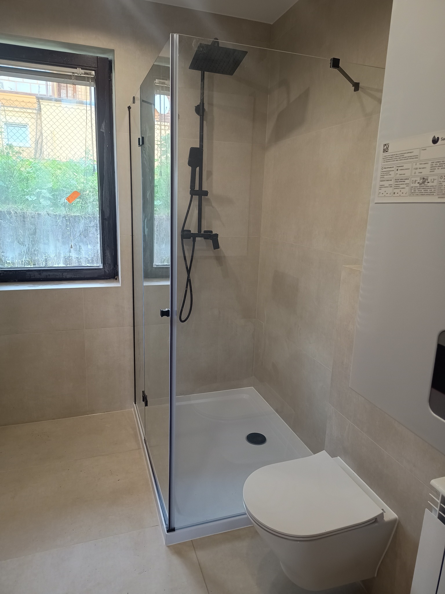 Wykończona łazienka z kabiną prysznicową typu walk-in, podwieszaną toaletą i beżowymi płytkami. Widoczne czarne elementy armatury i okno z widokiem na zieleń.