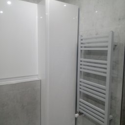 Prywatne - Biała szafka łazienkowa wisząca obok grzejnika drabinkowego na szarej ścianie imitującej beton.