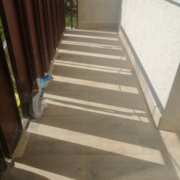 Prywatne - Wyłożony jasnymi płytkami balkon z brązową metalową balustradą, na podłodze leży zużyte opakowanie po produkcie budowlanym.
