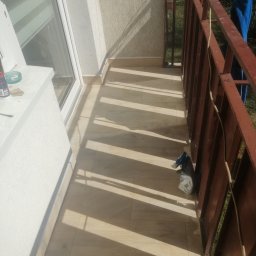 Prywatne - Wykończony balkon z brązową barierką, jasnymi płytkami podłogowymi i promieniami słońca tworzącymi geometryczne wzory, widoczny worek z materiałem budowlanym przy barierce.