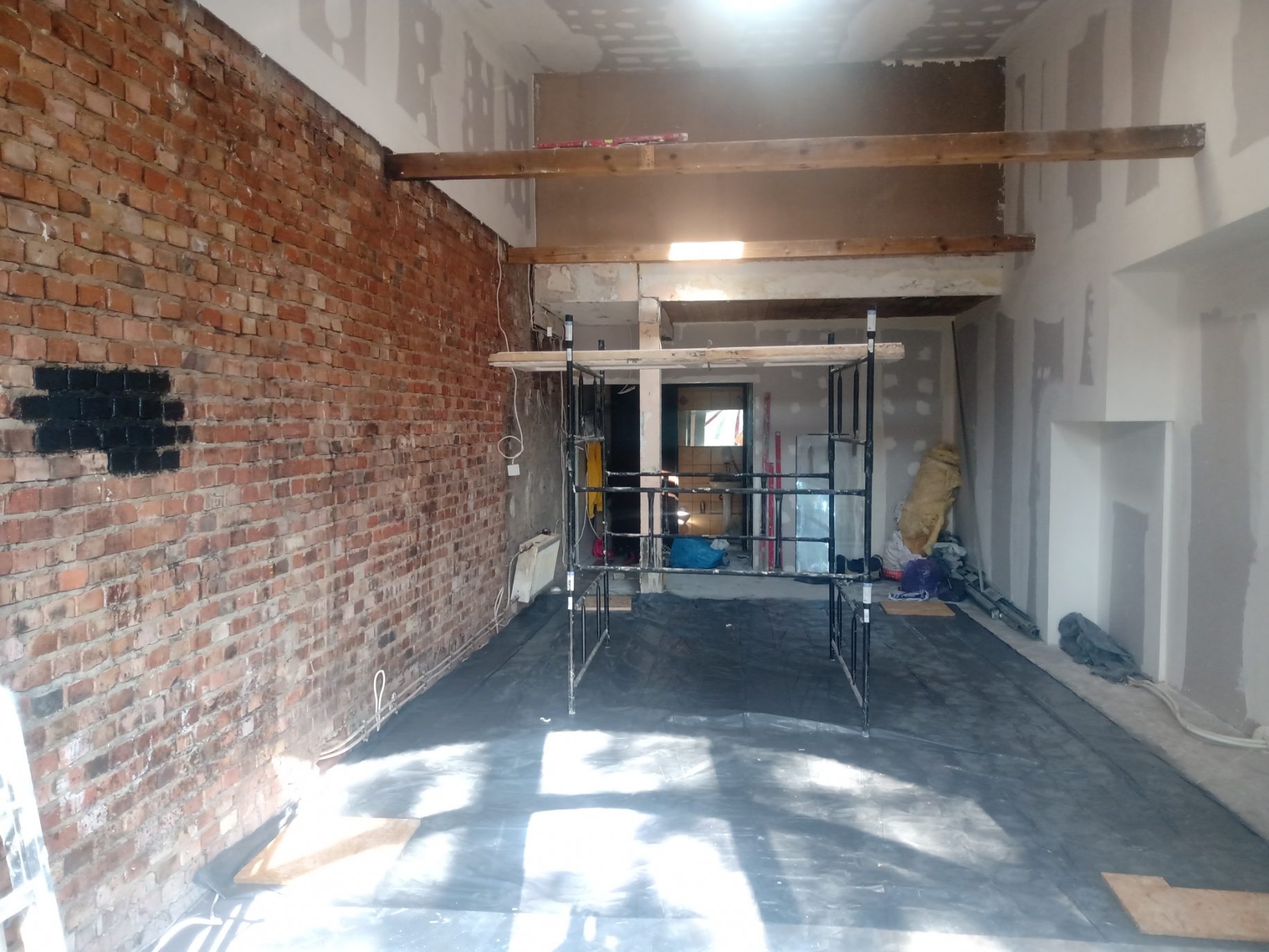 Wnętrze w trakcie remontu: surowa ceglana ściana z czarnymi fragmentami, rusztowanie, przygotowane ściany do szpachlowania, zabezpieczona podłoga folią, drewniane belki stropowe.