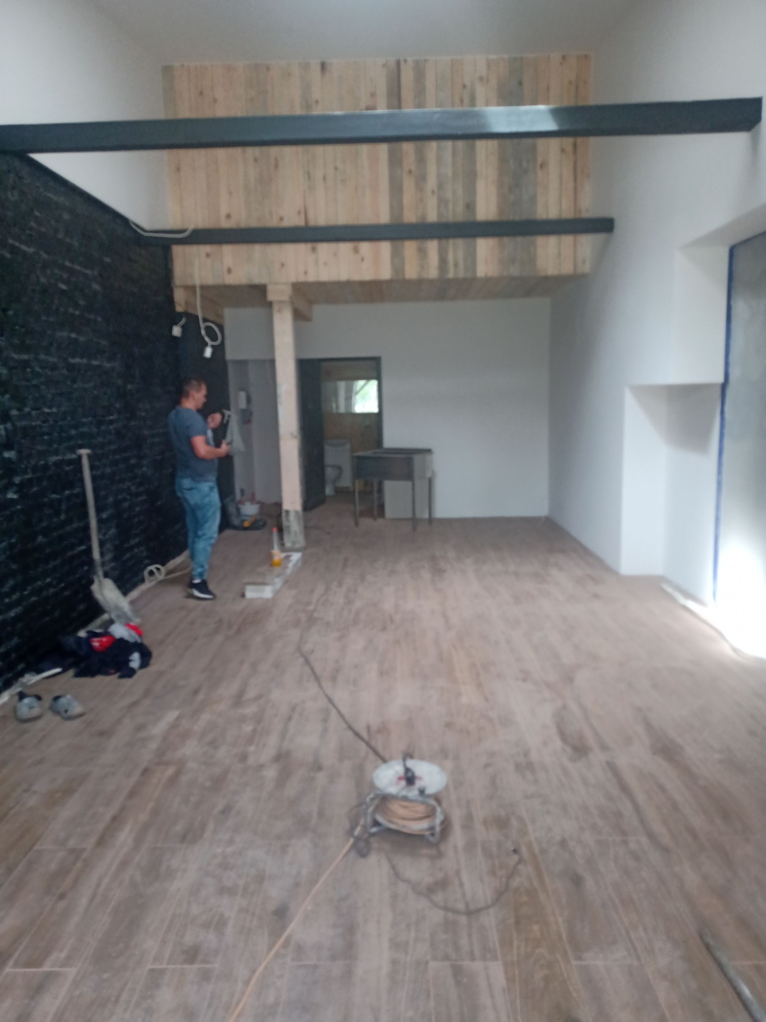 Wnętrze remontowanego pomieszczenia z drewnianą antresolą, widoczne narzędzia i materiały budowlane, podłoga wyłożona płytkami imitującymi drewno, ściana z czarnej cegły.