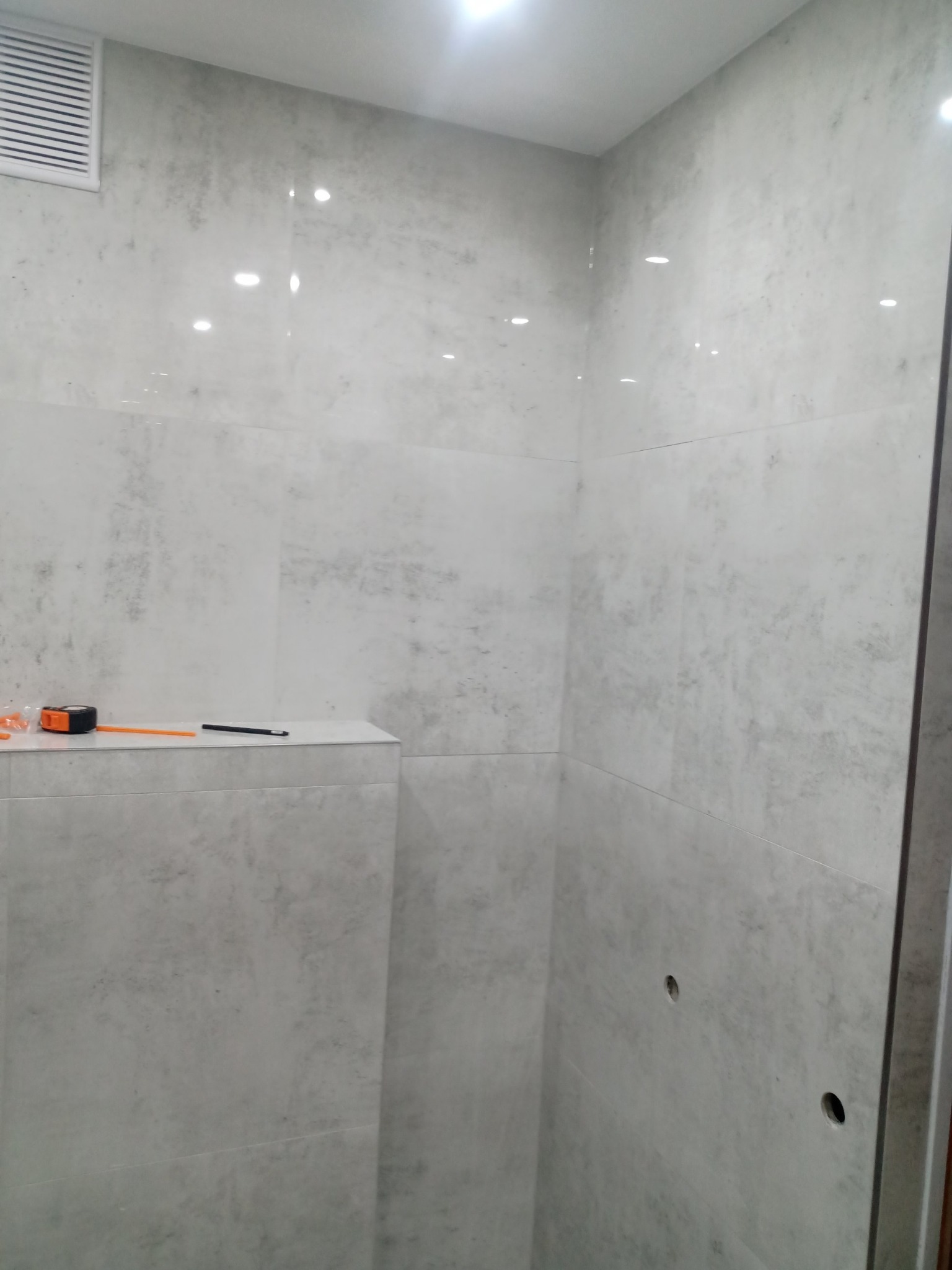 Świeżo wykafelkowana łazienka z szarymi płytkami imitującymi beton; na półce leży miarka i ołówek.