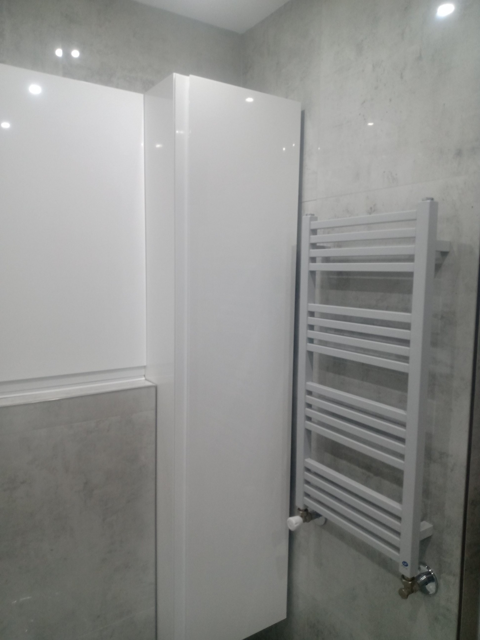 Biała szafka łazienkowa wisząca obok grzejnika drabinkowego na szarej ścianie imitującej beton.