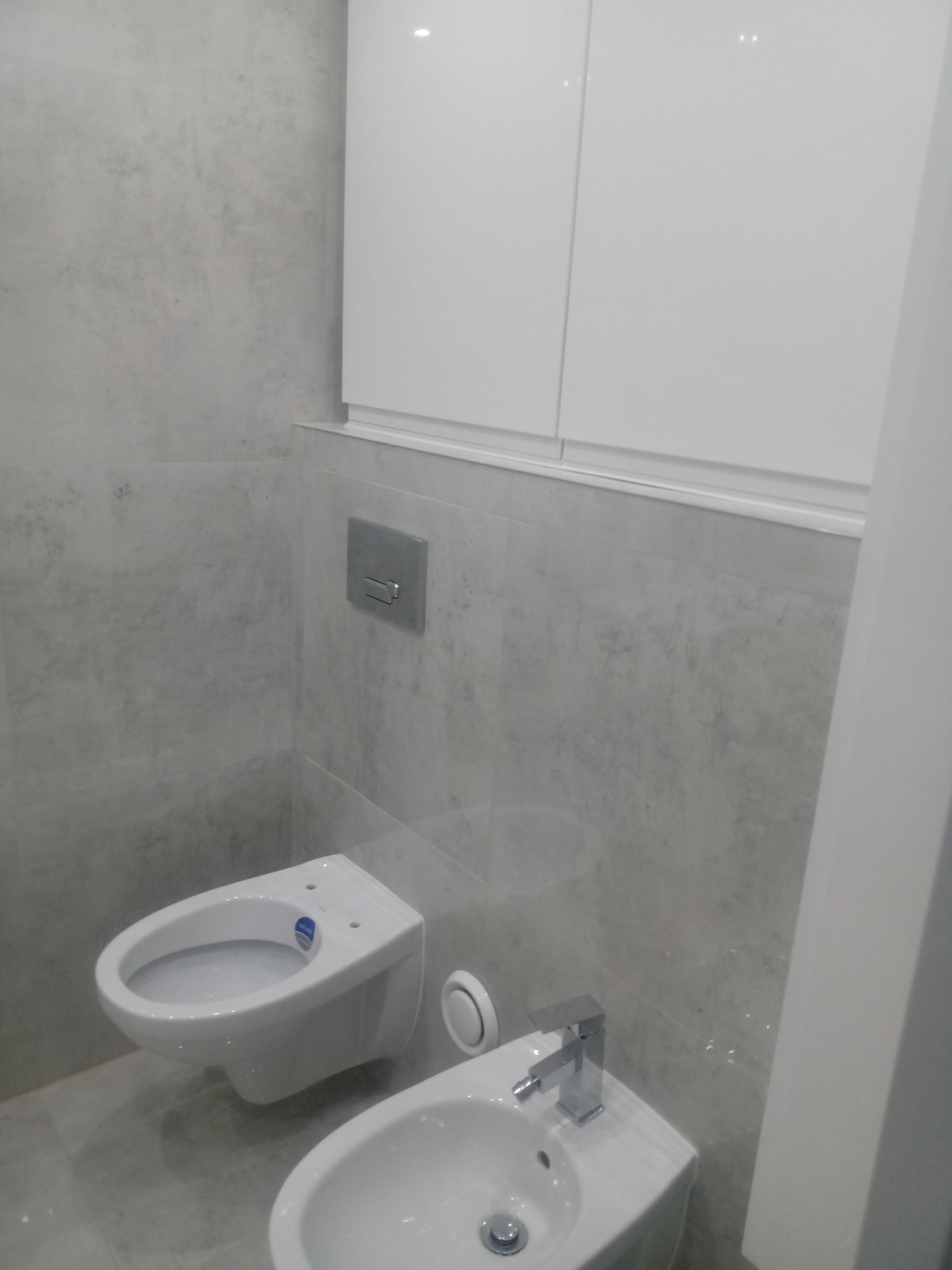 Nowoczesna łazienka z szarymi płytkami imitującymi beton, podwieszaną toaletą bez deski, bidetem z chromowaną baterią i białą szafką wiszącą.