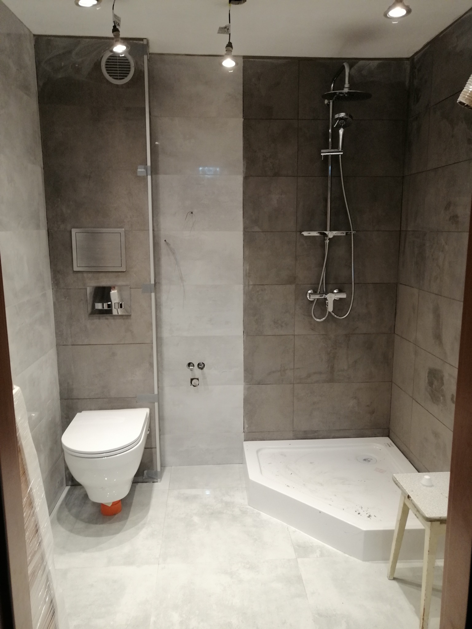 Nowoczesna łazienka z szarymi płytkami, białą toaletą z pomarańczowym elementem montażowym, kabiną prysznicową i minimalistycznym stołkiem.