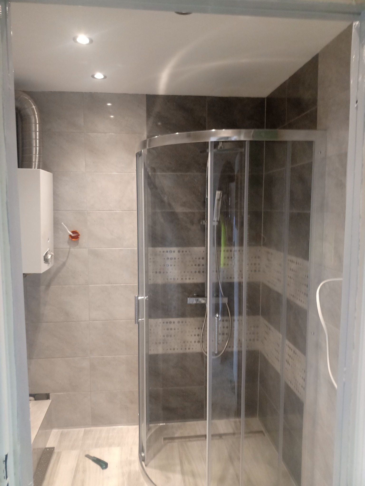 Nowoczesna kabina prysznicowa z zaokrąglonymi szybami w łazience wykończonej szarymi płytkami, widoczny bojler na ścianie i nóż na podłodze.