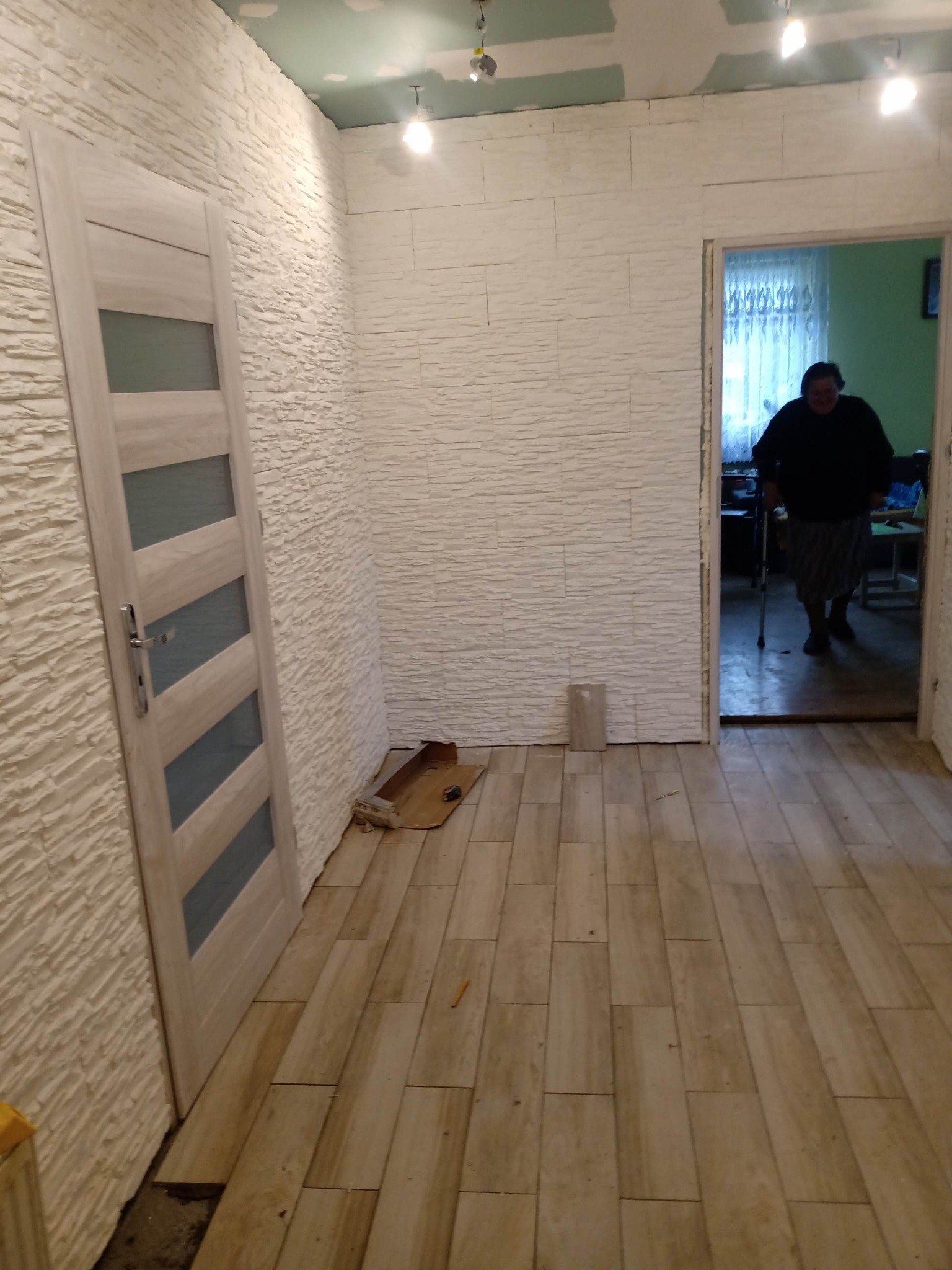 Wnętrze w trakcie prac wykończeniowych: ściana z dekoracyjną okładziną imitującą kamień, częściowo wykończony sufit, ułożona podłoga z płytek imitujących drewno, widoczna osoba w głębi pomieszczenia.