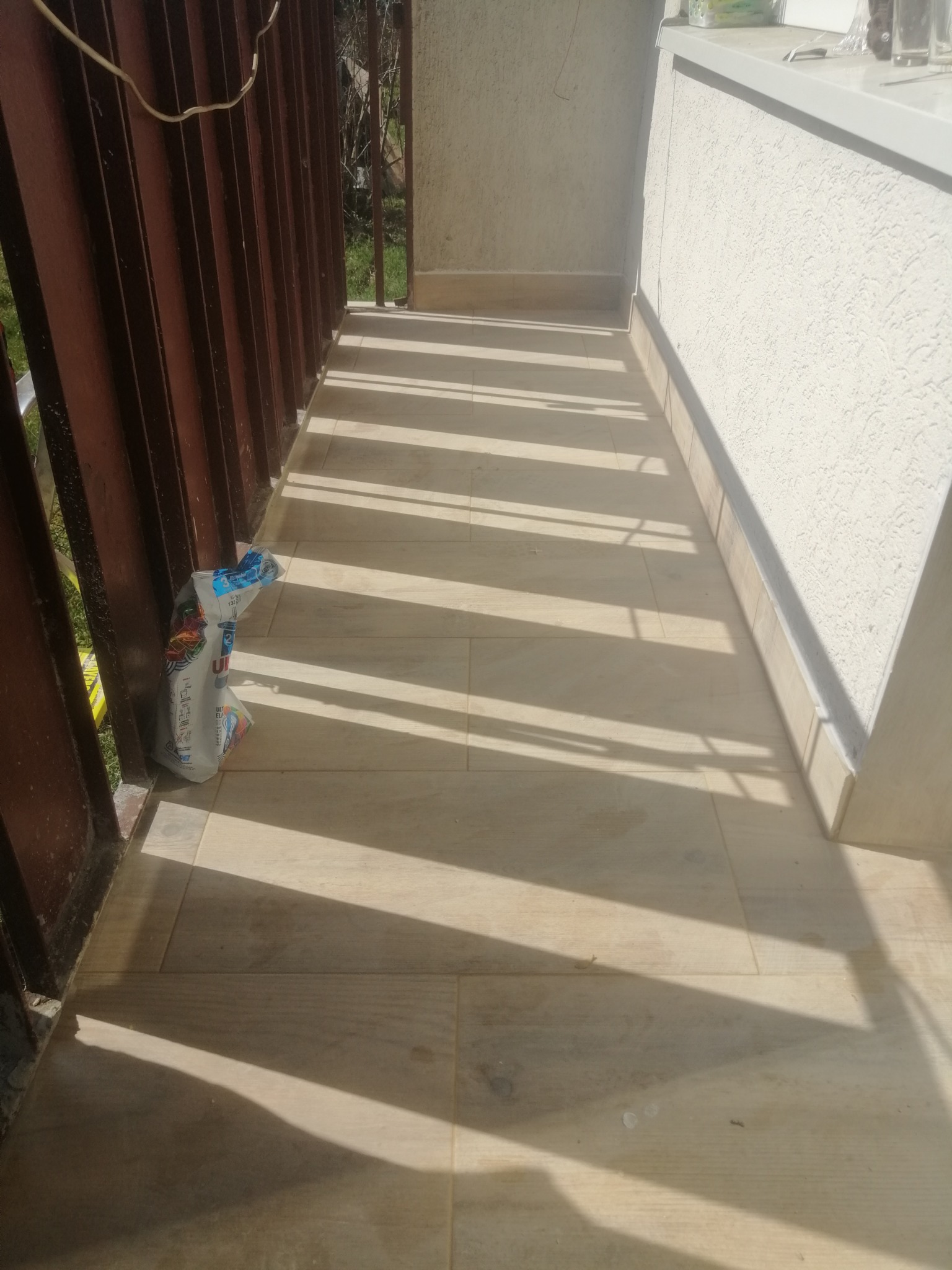 Wyłożony jasnymi płytkami balkon z brązową metalową balustradą, na podłodze leży zużyte opakowanie po produkcie budowlanym.