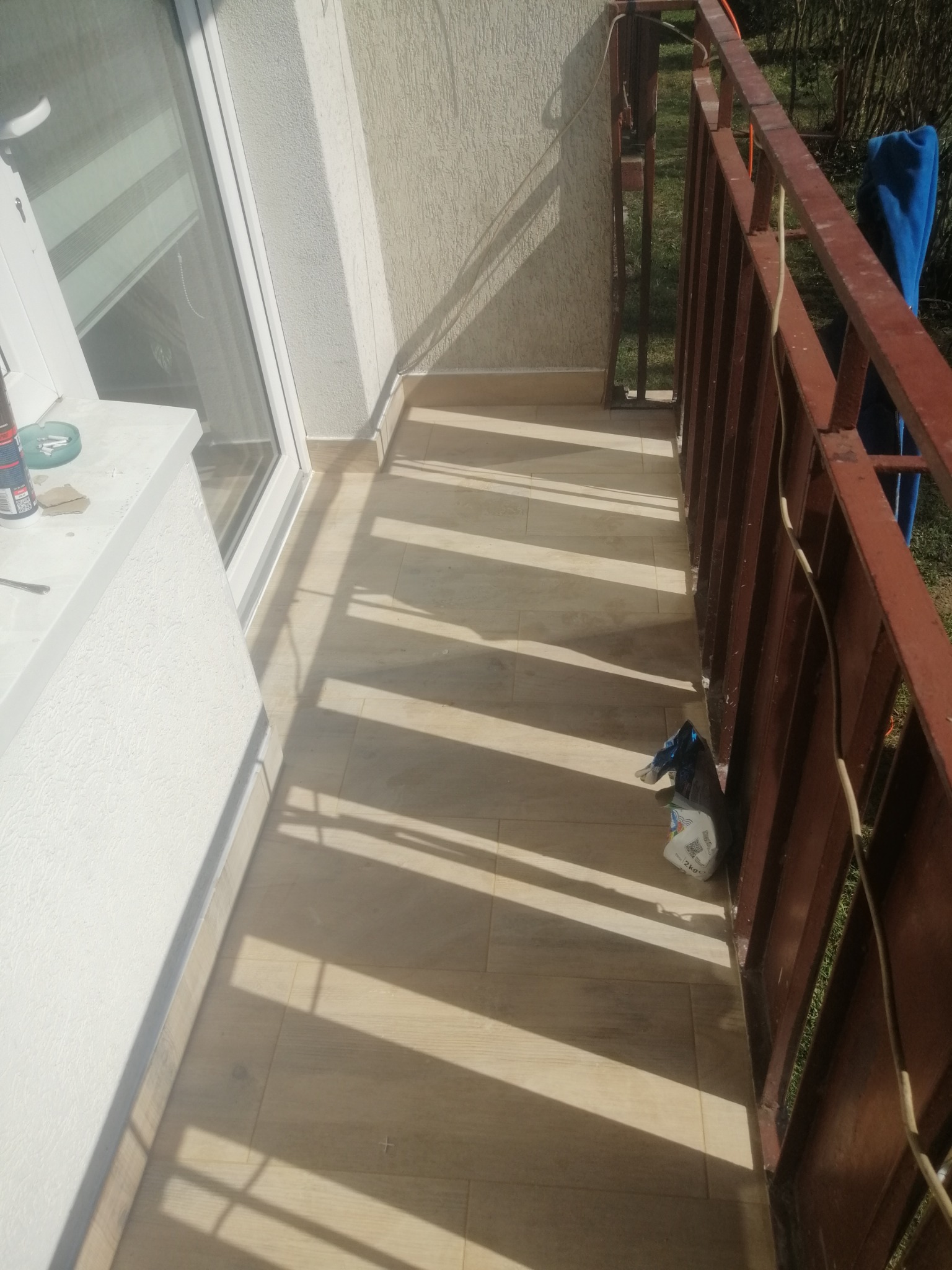 Wykończony balkon z brązową barierką, jasnymi płytkami podłogowymi i promieniami słońca tworzącymi geometryczne wzory, widoczny worek z materiałem budowlanym przy barierce.